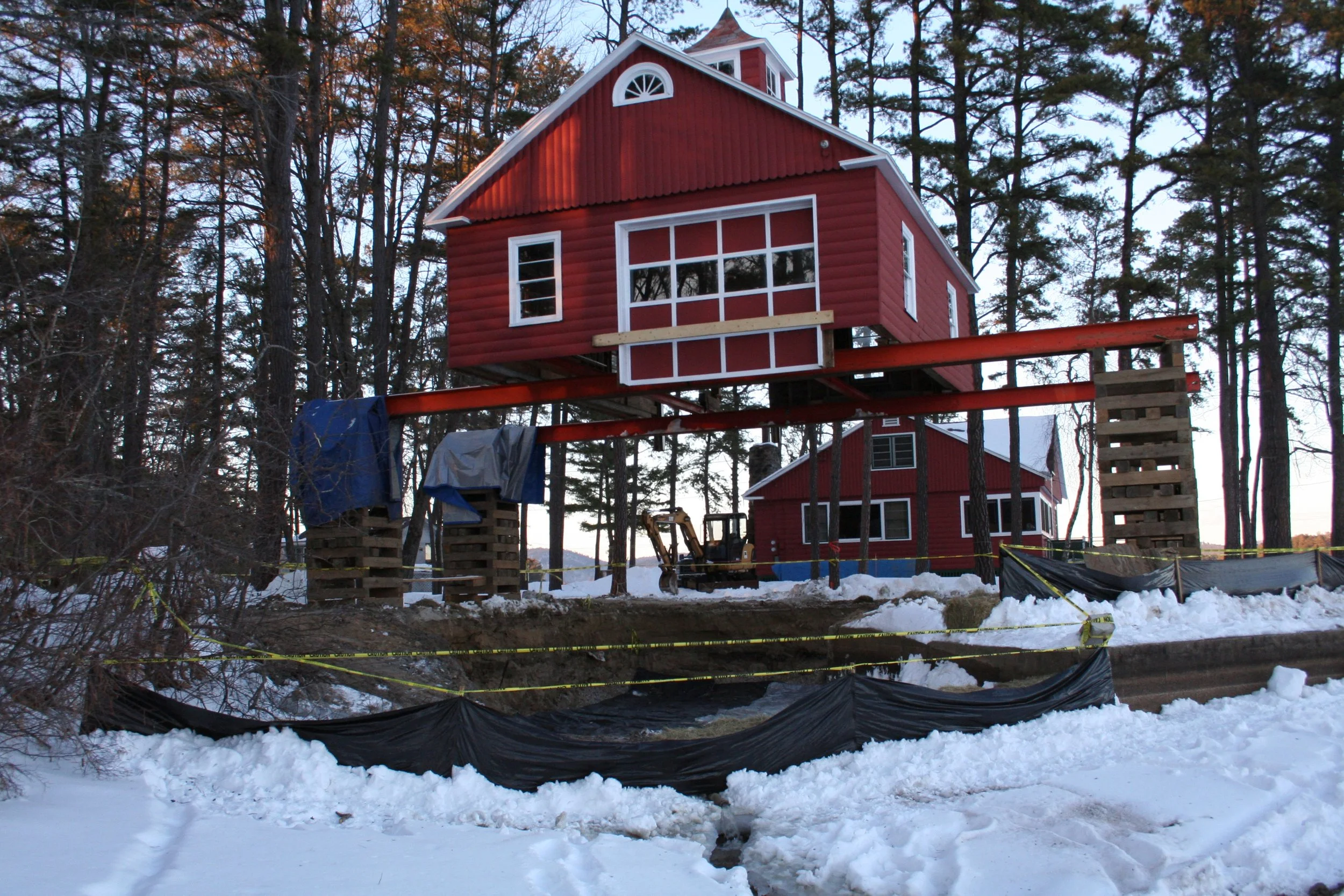 A house under construction with the upper part suspended by supports, surrounded by snow and construction barriers in a wooded area.