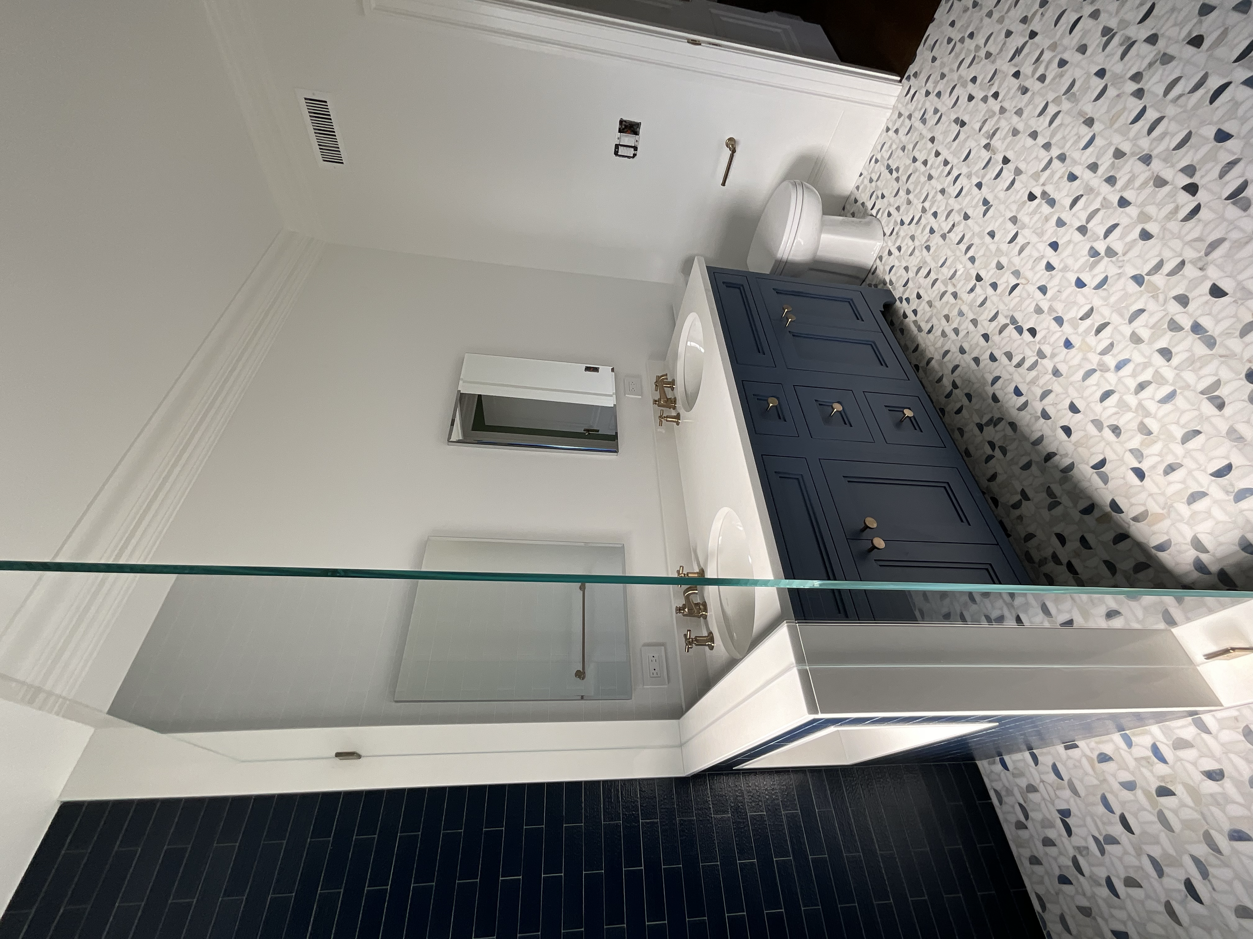 Modern bathroom with white walls and a navy blue vanity with gold hardware, double sinks, and a large mirror. There is a white toilet, patterned tile flooring, a glass shower enclosure, and a vent on the ceiling.