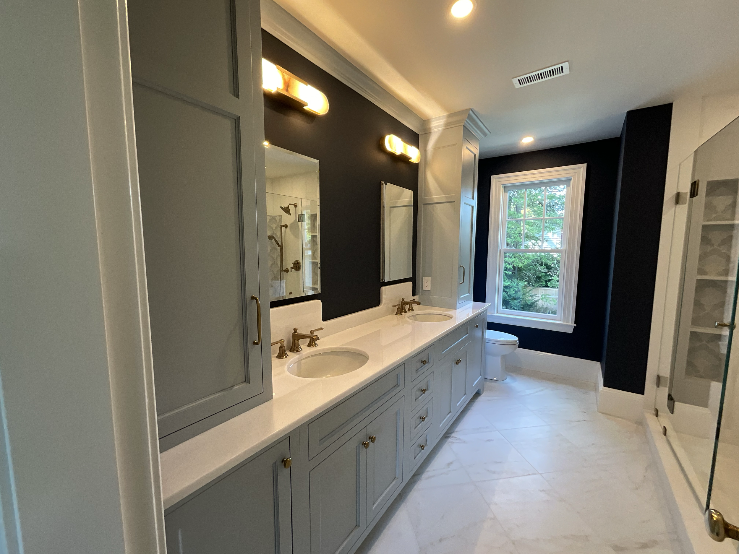 A modern bathroom with double vanity sinks, gold fixtures, dark accent wall, light gray cabinets, large window, and a glass shower enclosure.