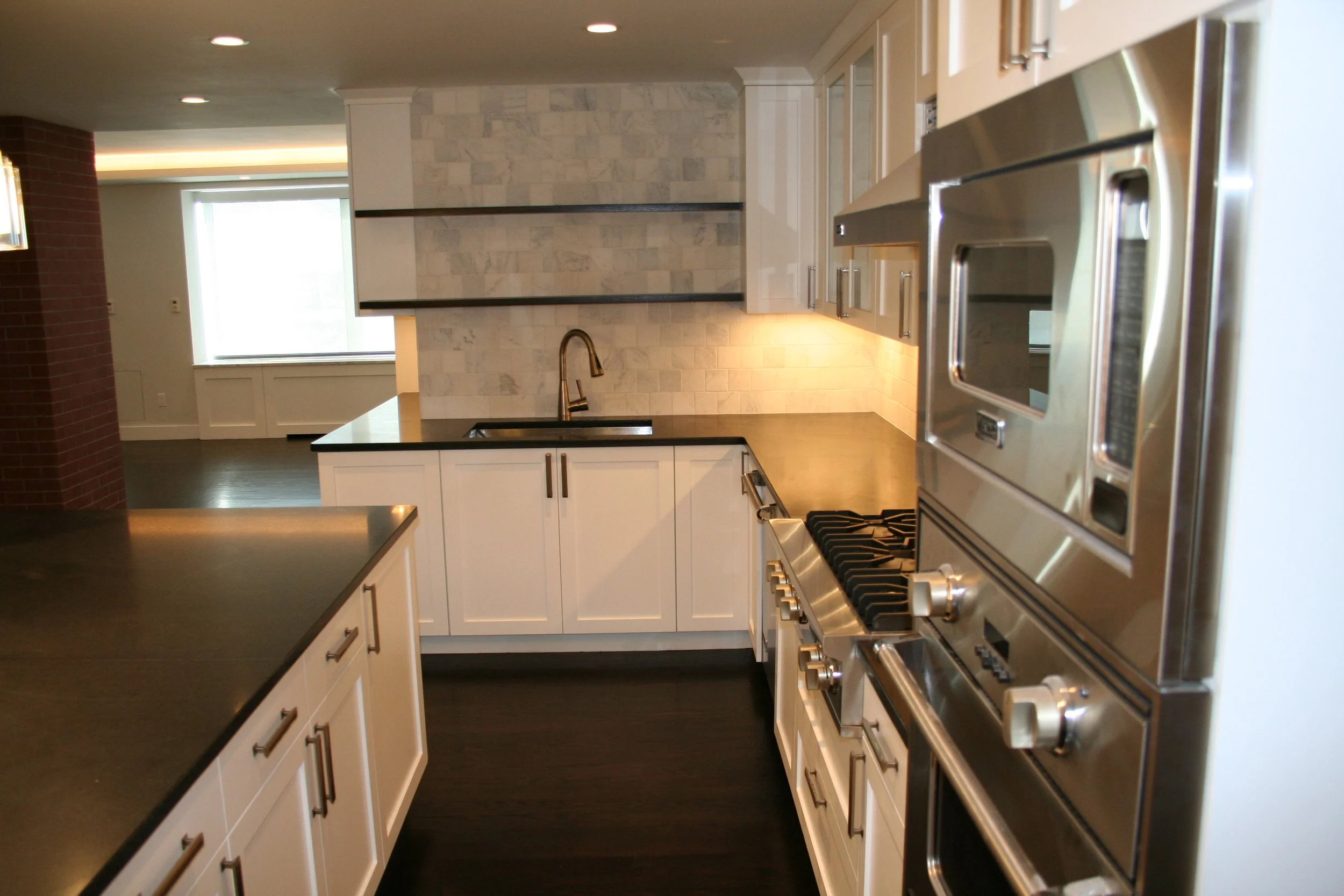 Modern kitchen with white cabinets, black countertops, stainless steel appliances, and a marble backsplash, with open shelving and a view into the living area.