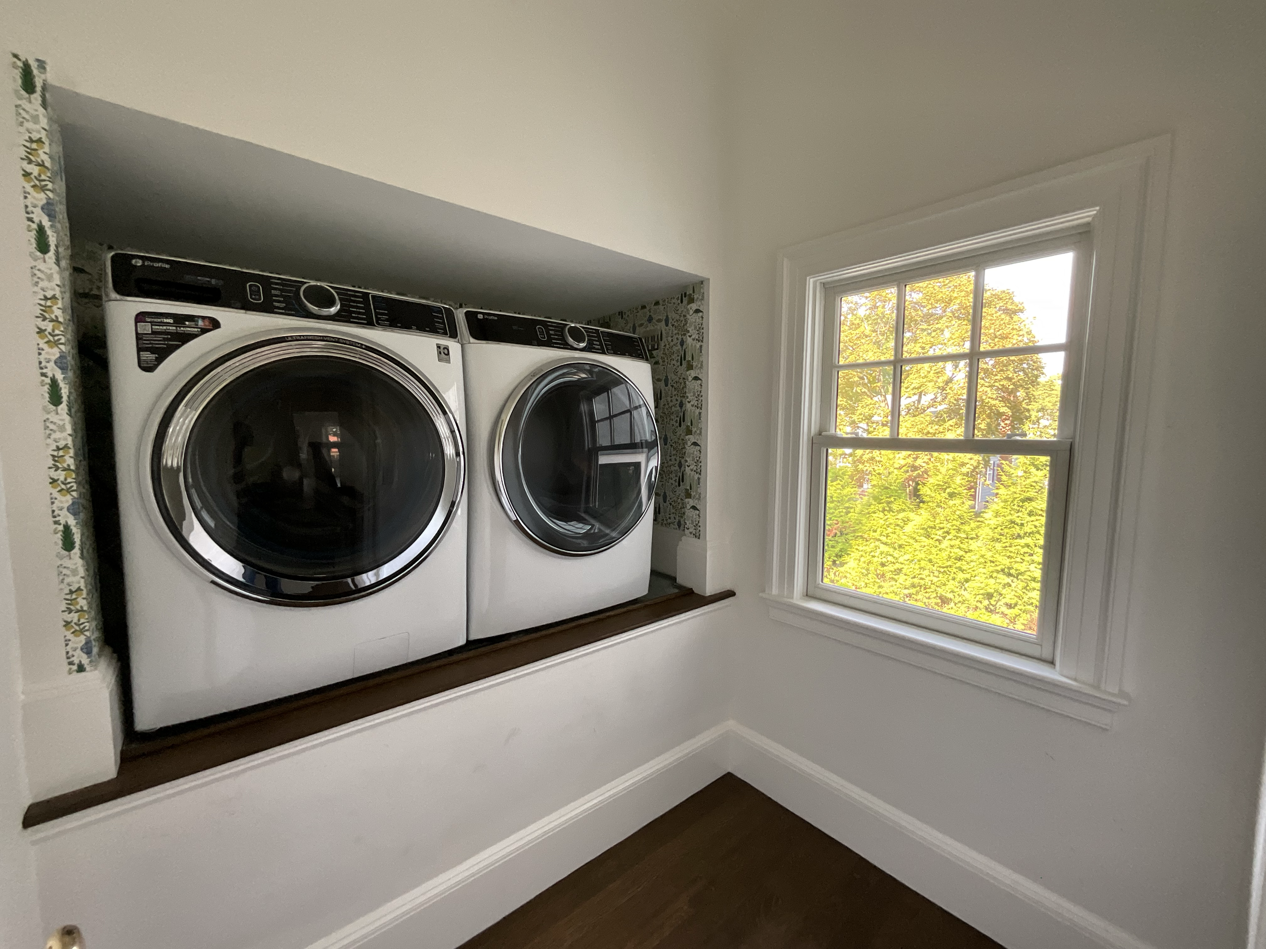 In-unit laundry room with a side-by-side washer and dryer, white walls, a window showing green trees outside, and a wooden shelf or ledge beneath the appliances.