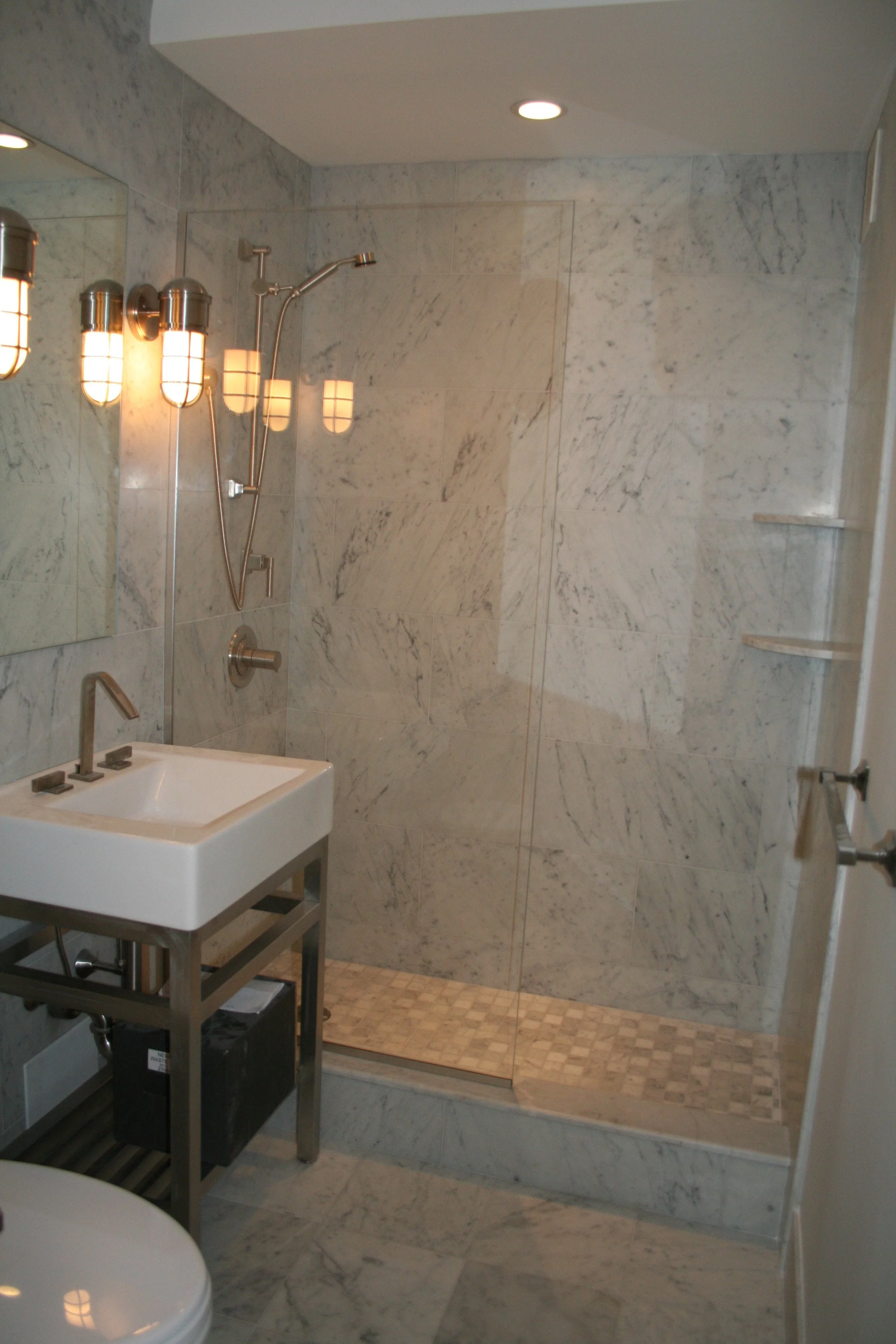 A modern bathroom with a walk-in shower featuring a glass door, marble walls, a small sink with a metal frame, and contemporary wall-mounted light fixtures.