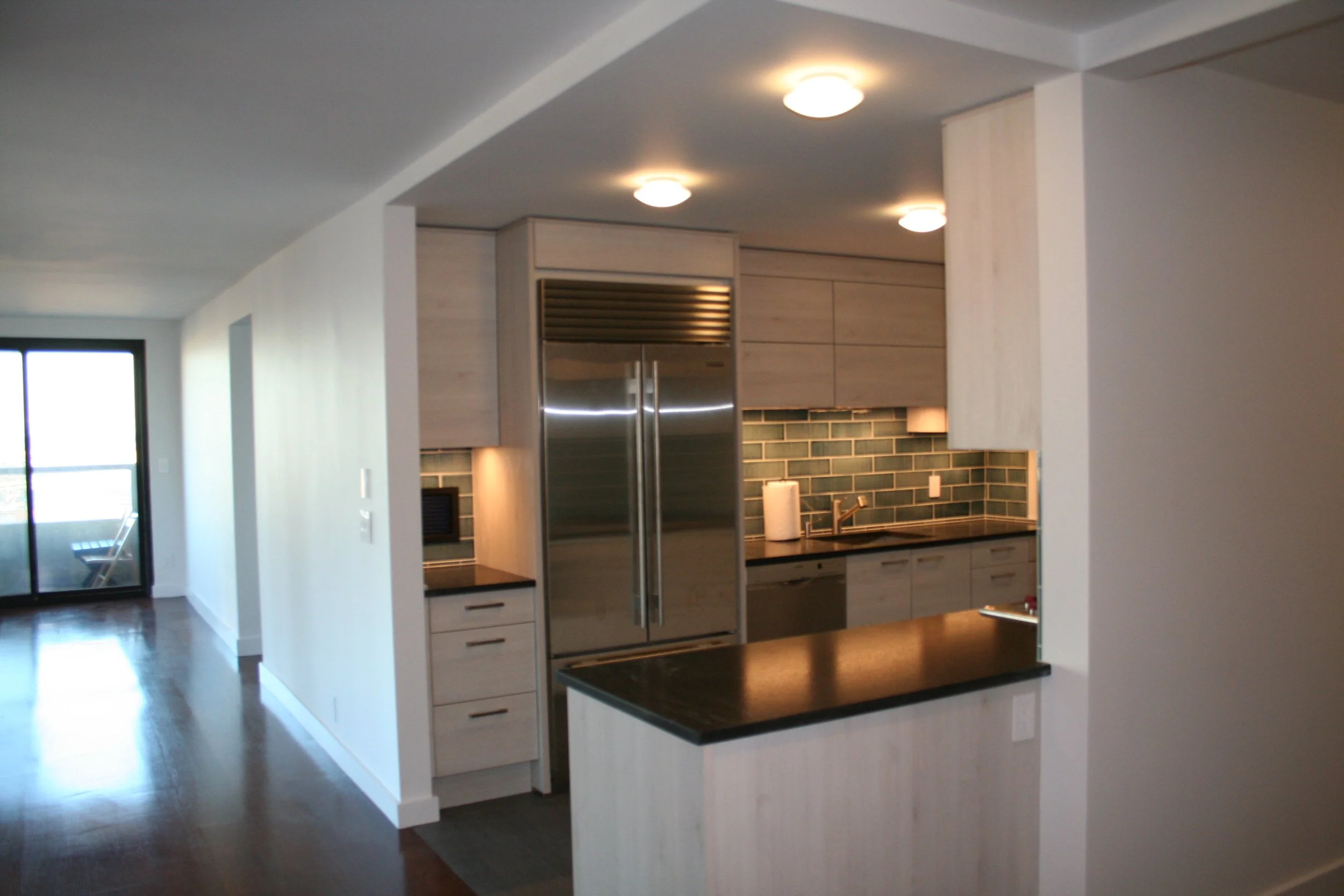 Modern kitchen with stainless steel refrigerator, dark countertop, green subway tile backsplash, and light wood cabinets.