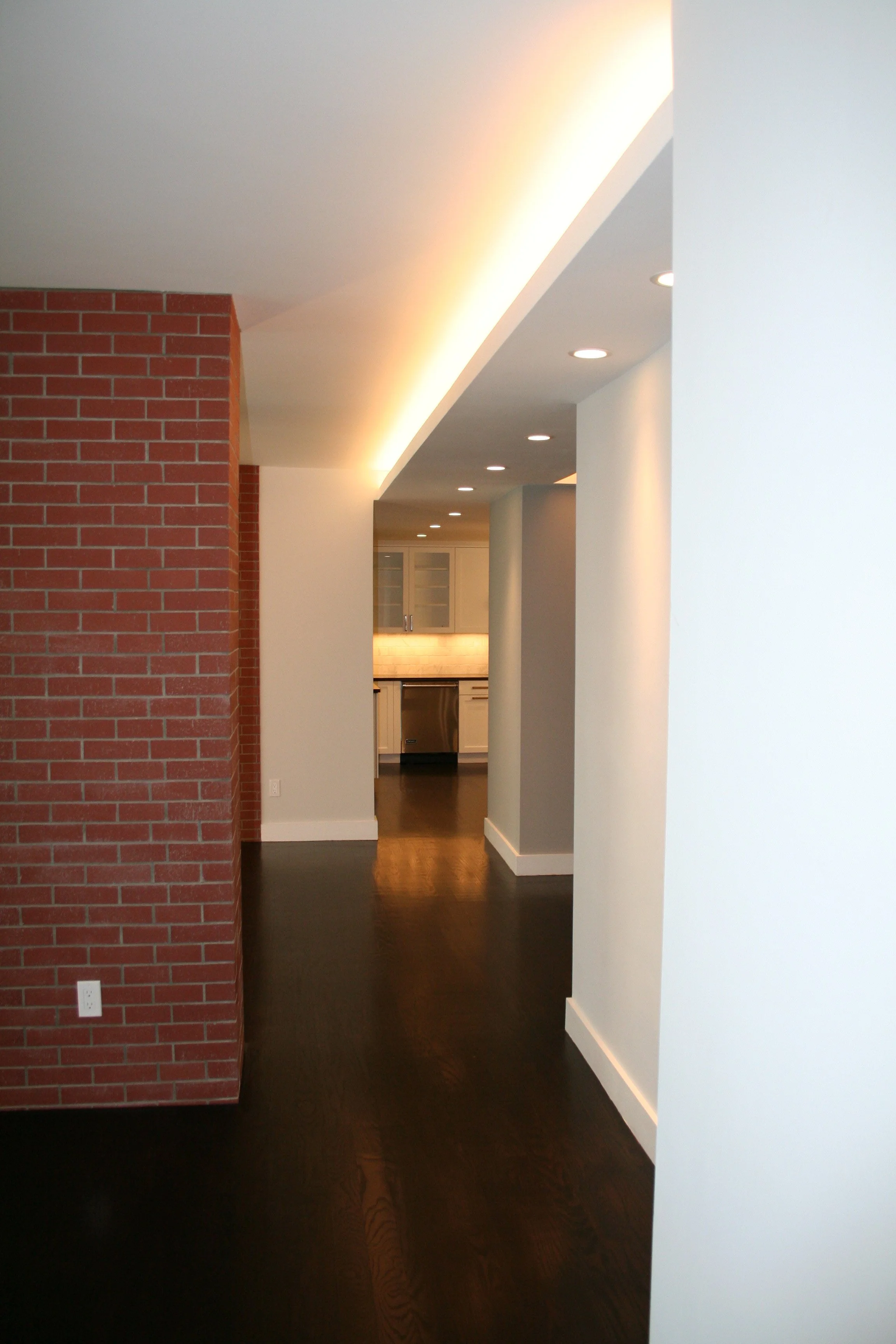 Interior view of a modern apartment with a dark wooden floor, white and gray walls, and a kitchen in the background. There is a red brick wall on the left.