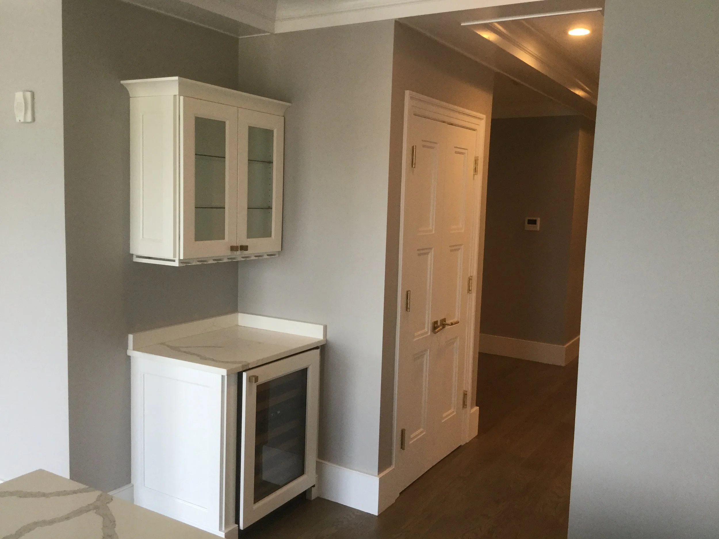 White kitchenette with upper glass-front cabinets and a mini wine fridge on the lower level, next to a white door in a neutral-colored room.