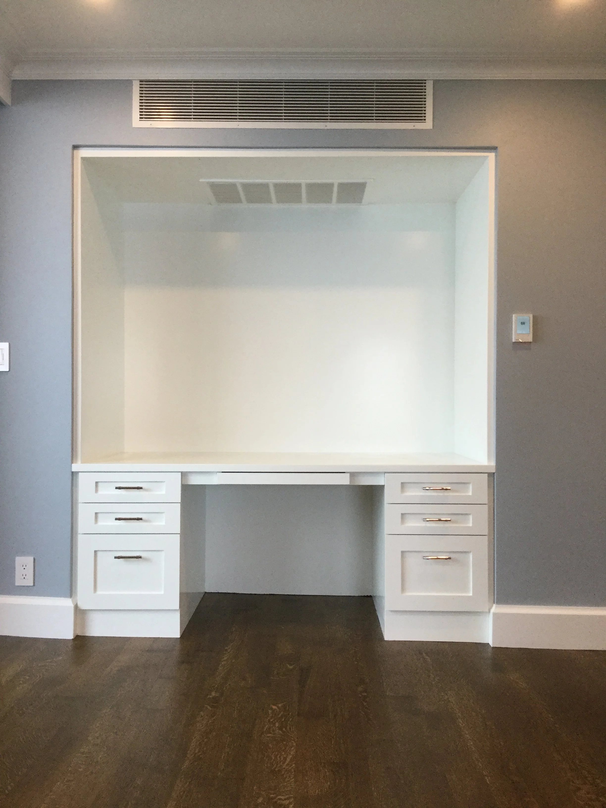 Empty built-in white desk with six drawers and a large open space in a room with light gray walls and dark hardwood floor.