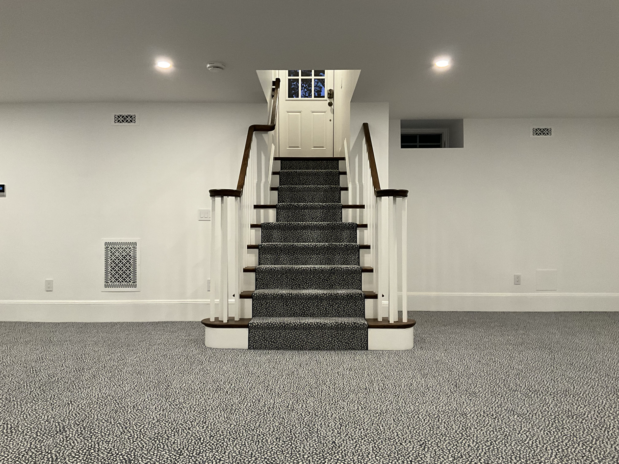 Interior view of a staircase with carpeted steps, white handrails, and a white door at the top, in a room with white walls, ceiling lights, and gray patterned carpet.