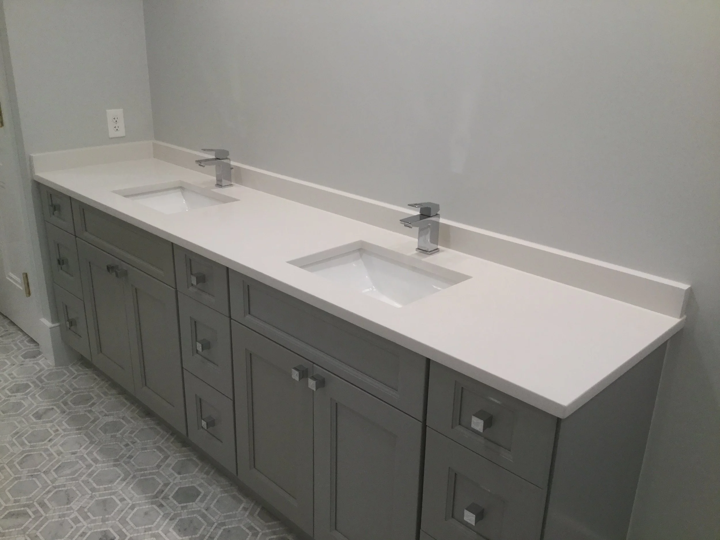 Double bathroom vanity with gray cabinetry, white countertop, and two rectangular sinks with modern chrome faucets, against a light gray wall, with patterned tile flooring.