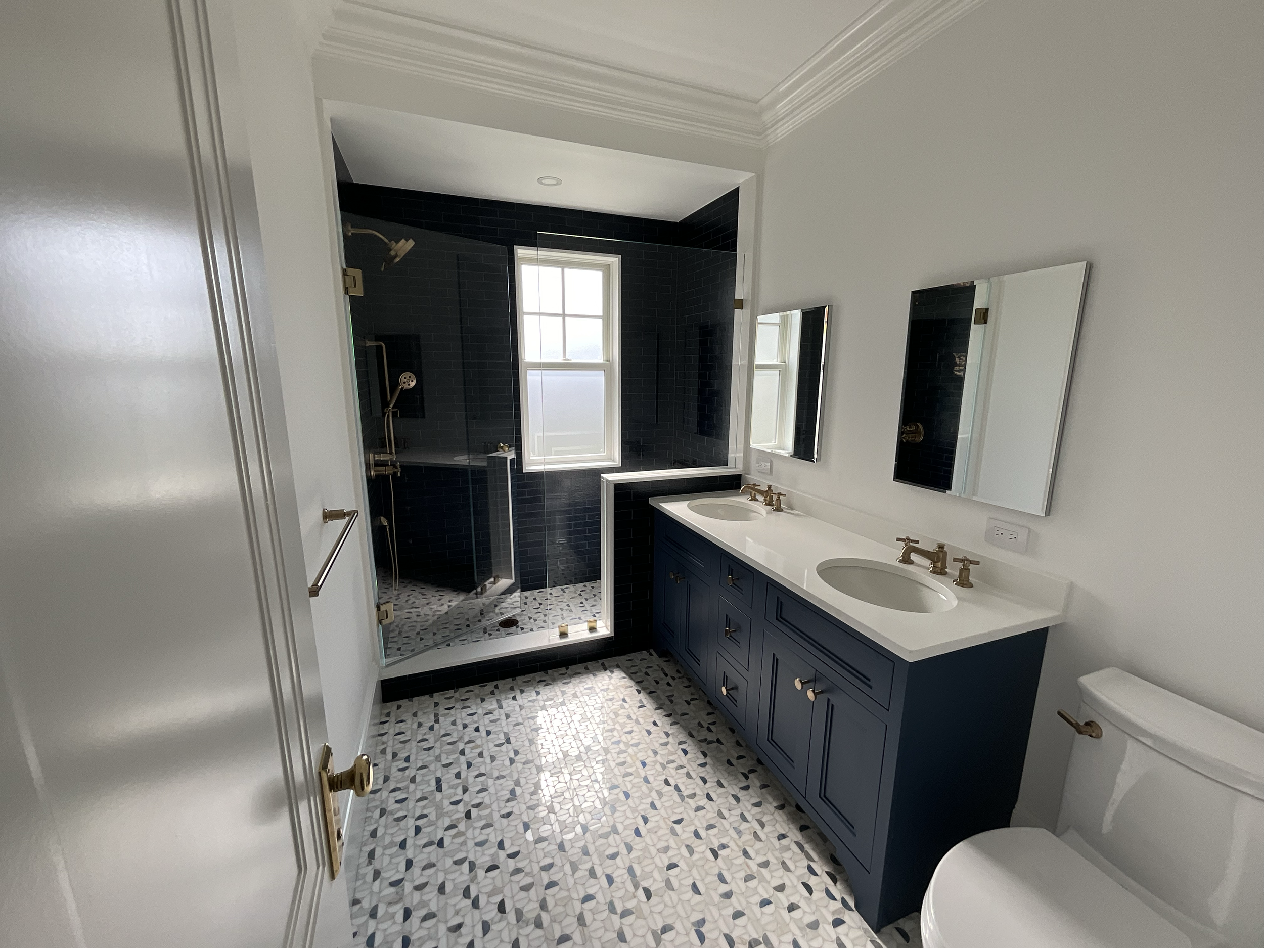 Modern bathroom with navy blue double vanity, white countertop, and two mirrors, adjacent to a walk-in shower with black tiles and pebble shower floor.