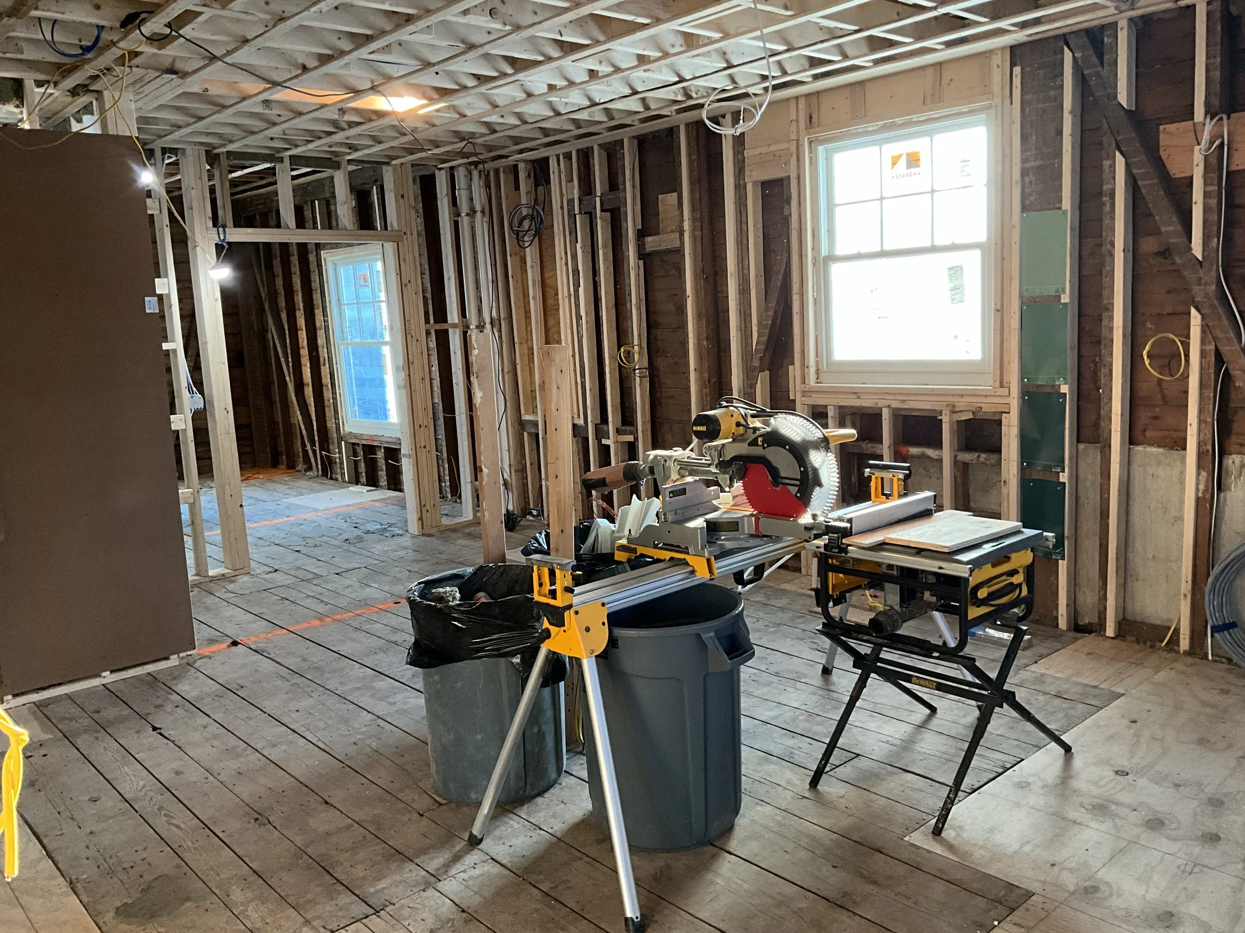 Interior of a house under construction with exposed wooden framing, two windows, and construction tools including a miter saw and a table saw.