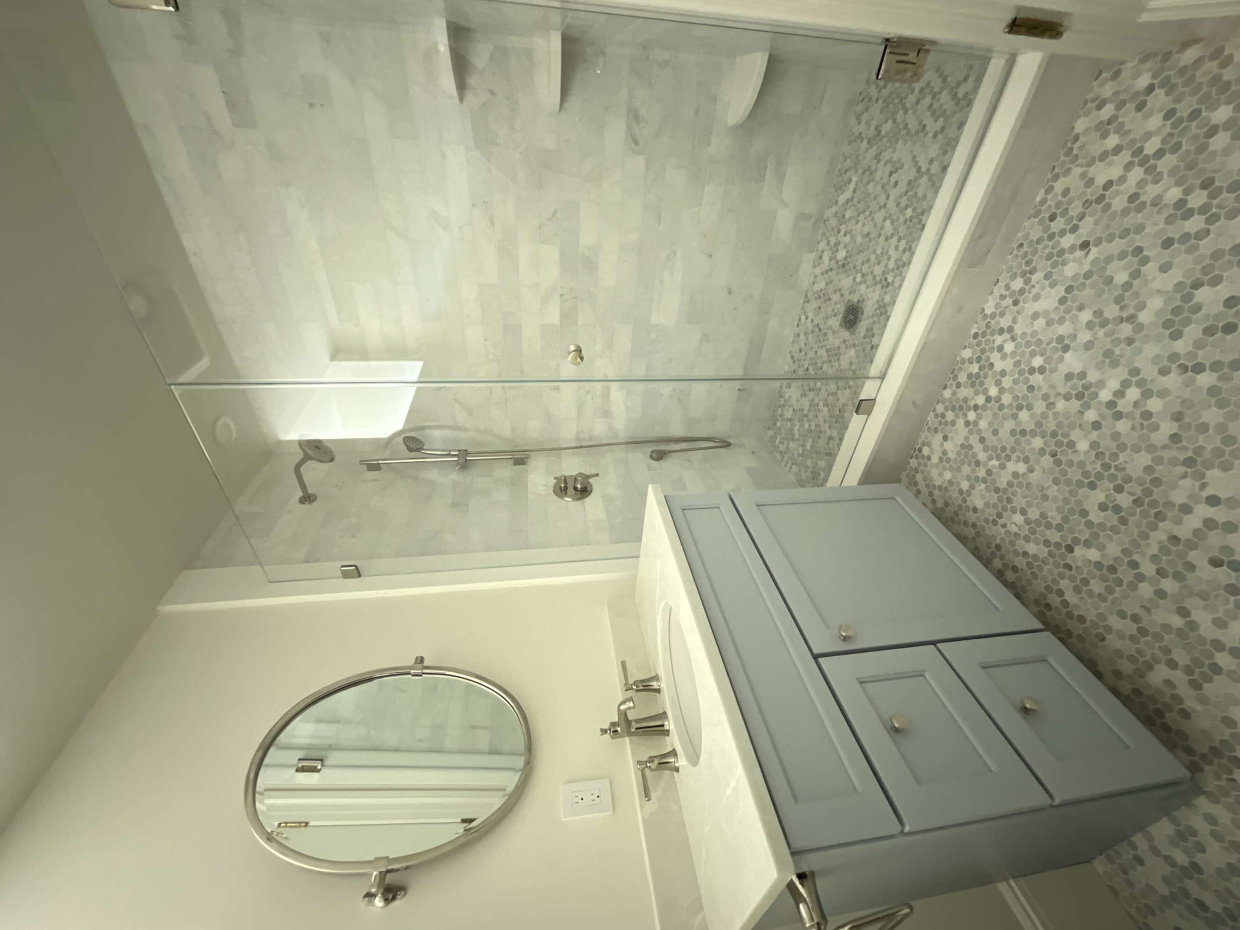 Bathroom with a glass-enclosed shower, a vanity with a round mirror, a sink with a metal faucet, and a light gray cabinet, with patterned floor tiles.