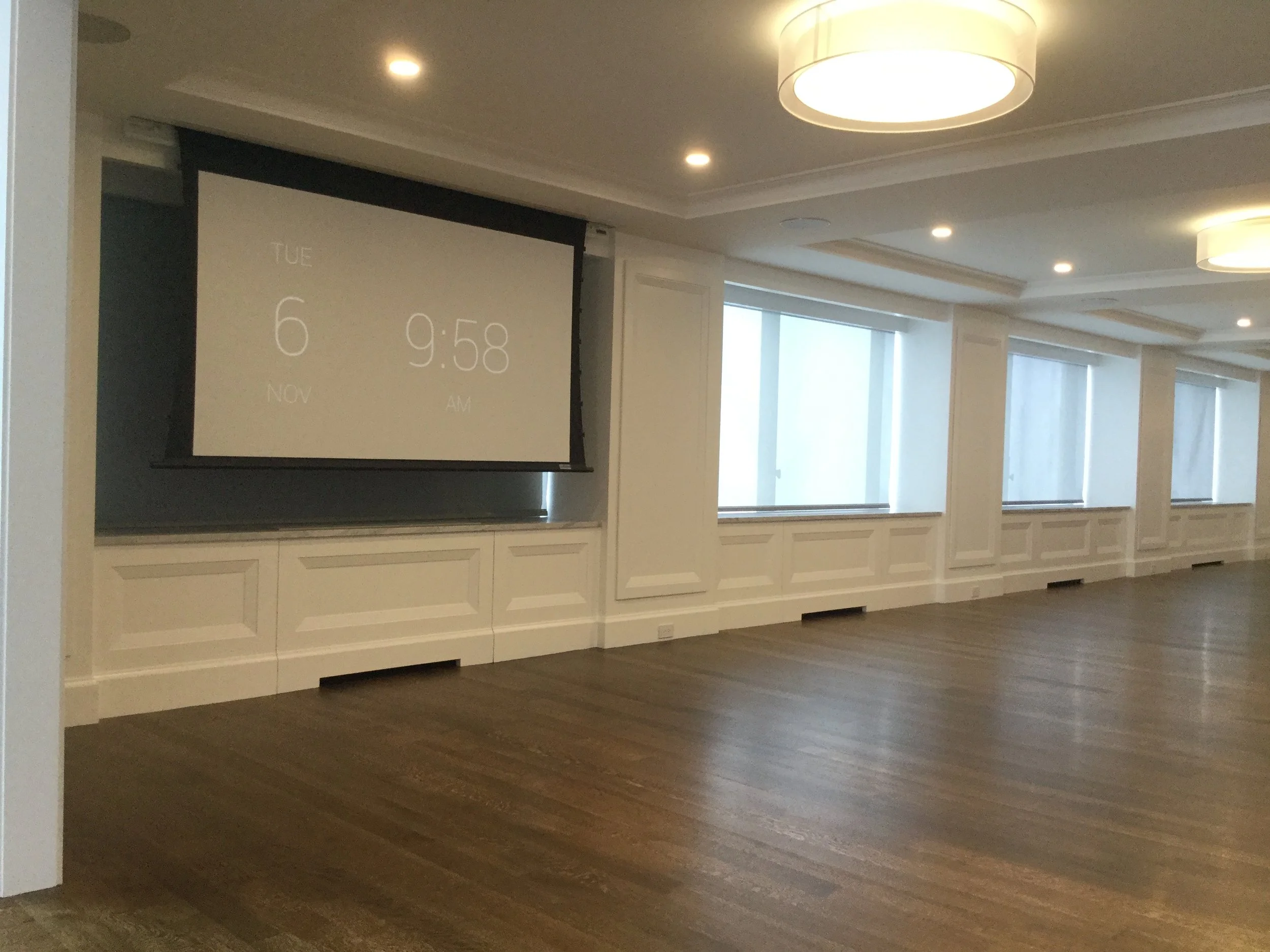 Empty conference room with wooden floors, white paneled walls, and large windows covered with shades. A projection screen displays the date and time: Tuesday, November 6 at 9:58 AM.