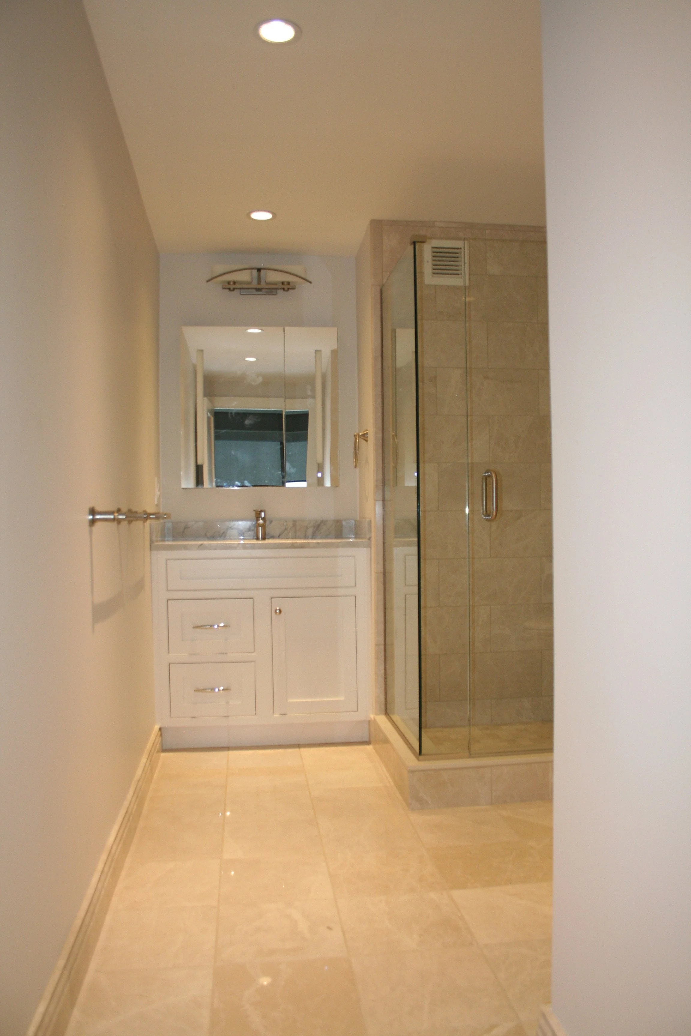 A bathroom with neutral-colored tiles, a white vanity with a marble top, a mirror, a glass-enclosed shower, and recessed ceiling lights.