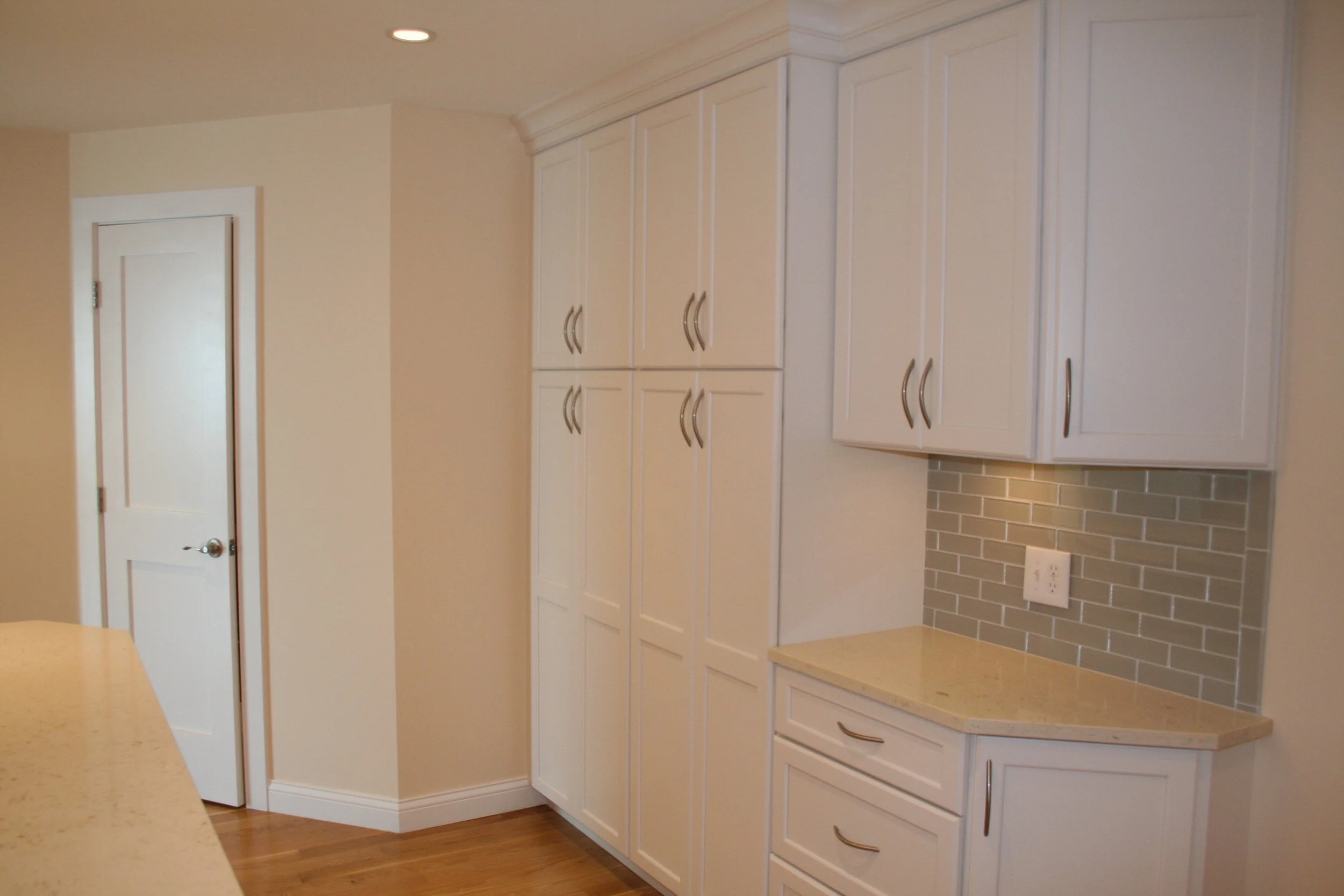 Kitchen with white cabinets, beige granite countertop, gray brick backsplash, electric outlet, corner of a beige countertop, and a closed white door.