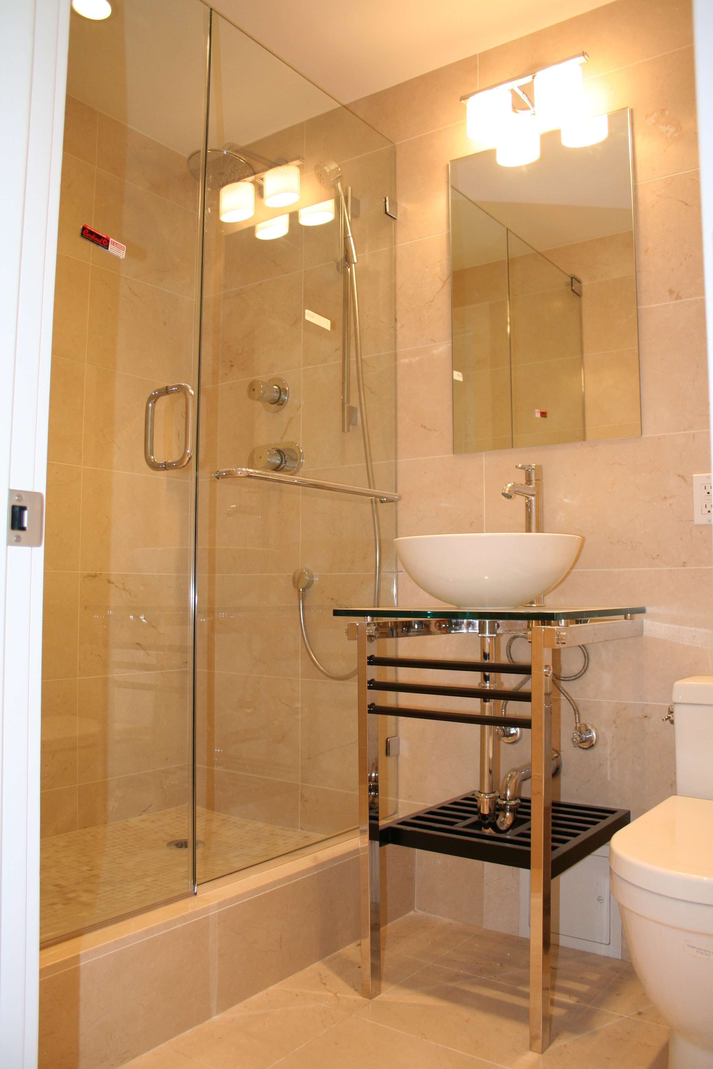 A modern bathroom with beige tiled walls and flooring, featuring a glass-enclosed shower with thermostatic controls, a white vessel sink on a metal and glass stand, a mirror above the sink, and a white toilet, all illuminated by overhead lighting.