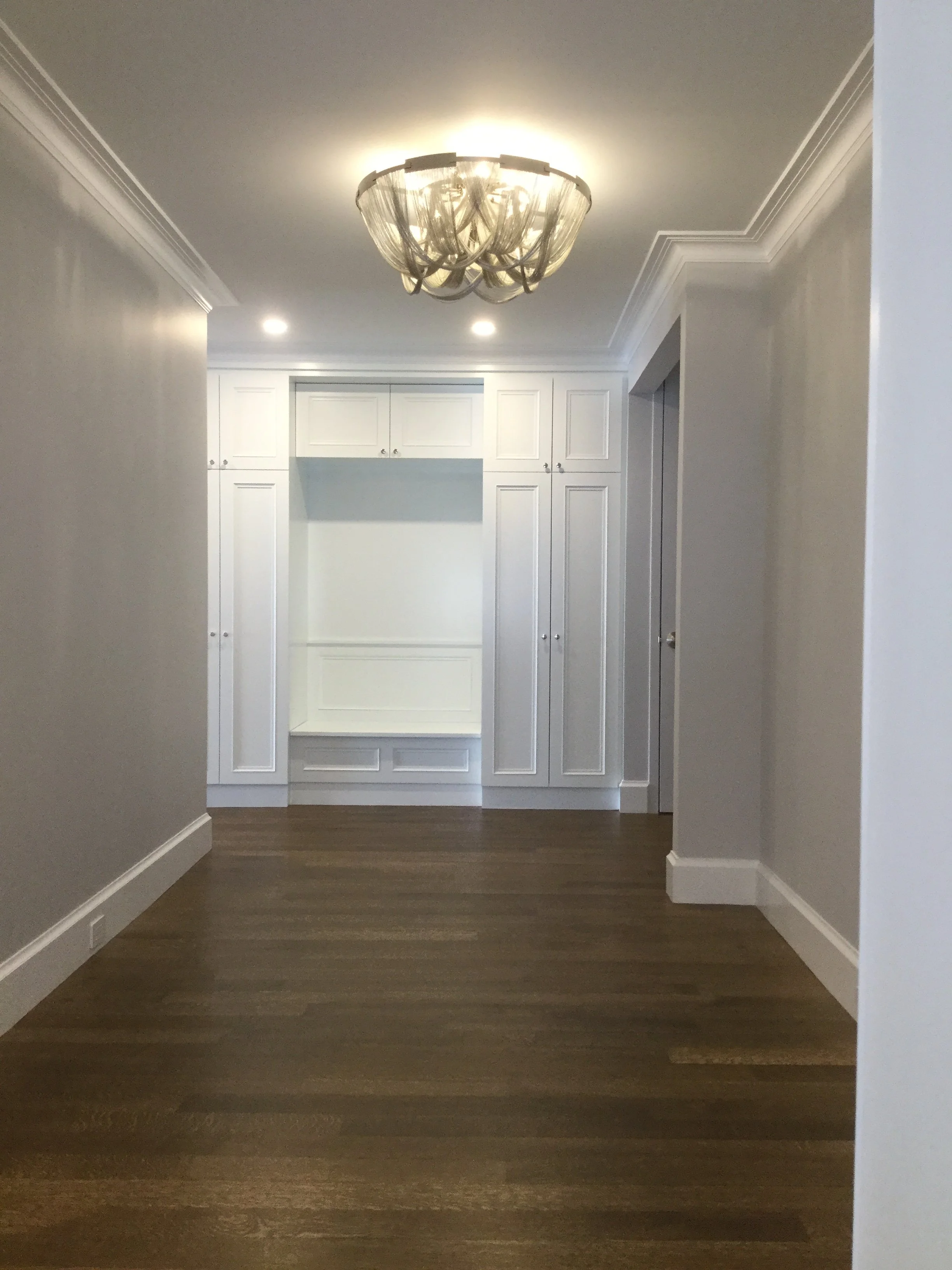Empty hallway with hardwood floors, white walls, inset paneling, and white built-in cabinets at the end. A central chandelier and two ceiling lights illuminate the space.