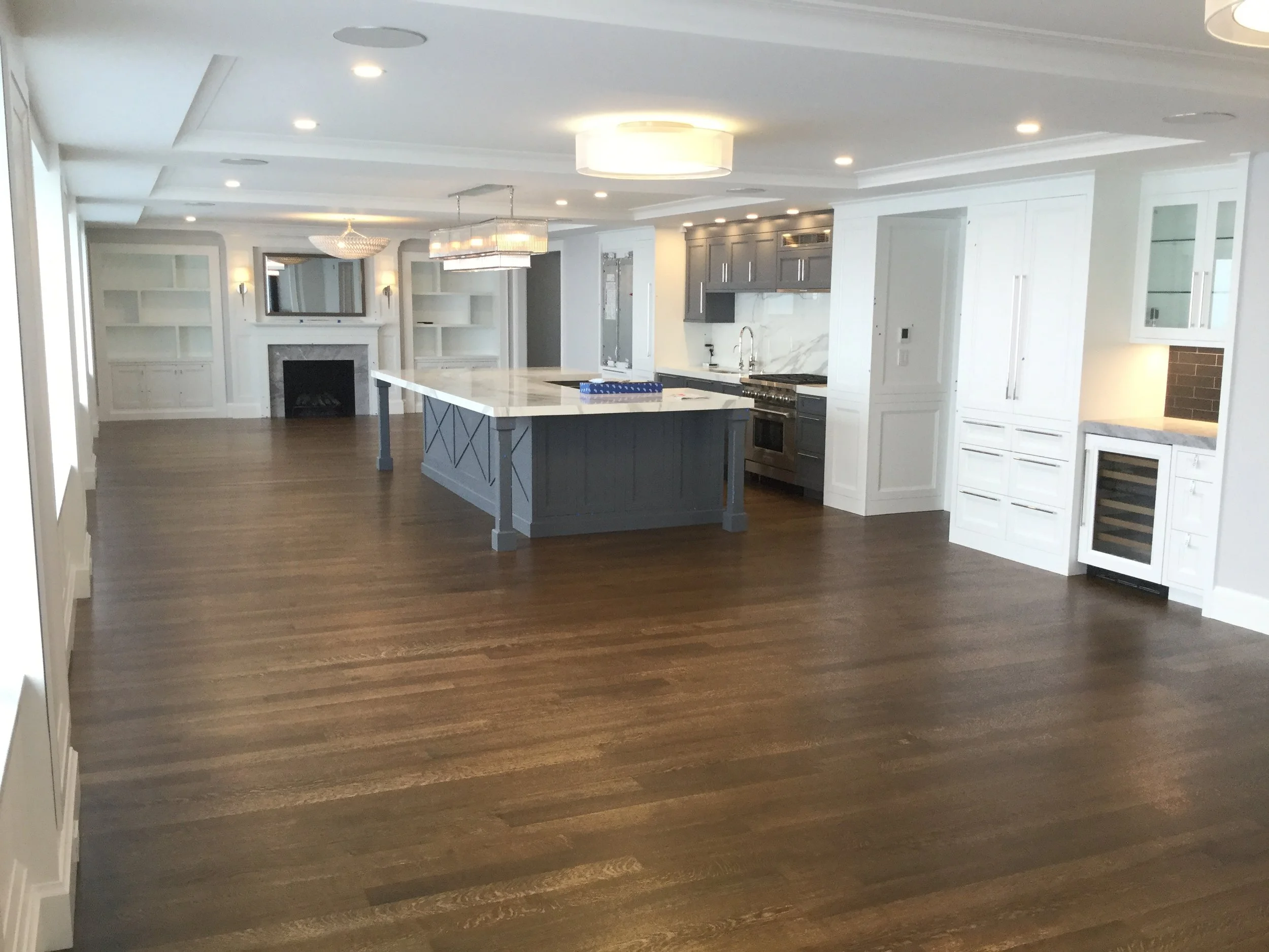 Open-concept kitchen and living room with dark hardwood floors, white walls, modern appliances, and a marble-clad island with a gray base in the center. There is a fireplace with shelving on either side and contemporary ceiling lighting fixtures.