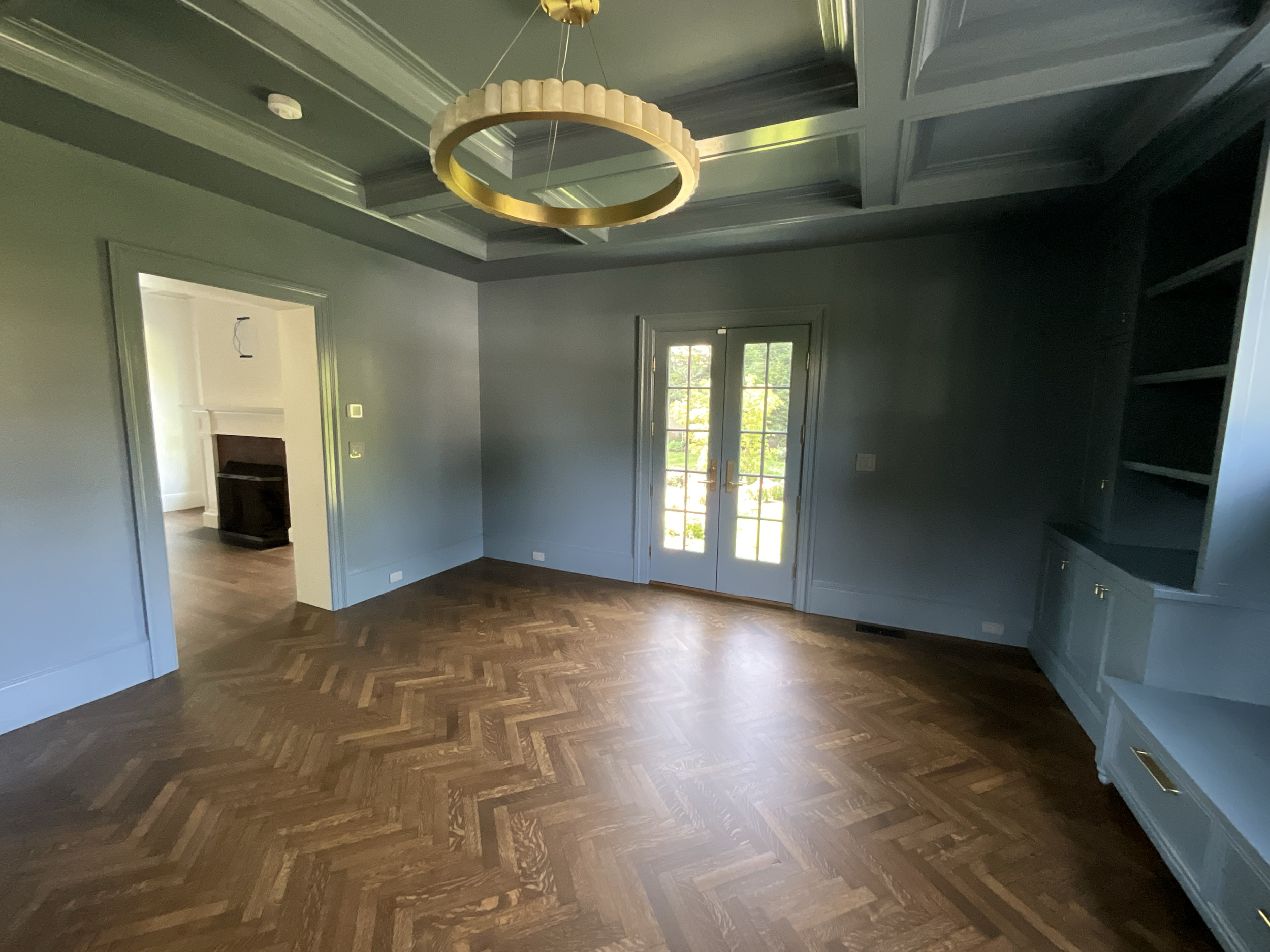 Empty living room with dark wood floor, blue walls and ceiling, built-in blue shelves, a double glass door opening to outside, and a gold circular light fixture on the ceiling.