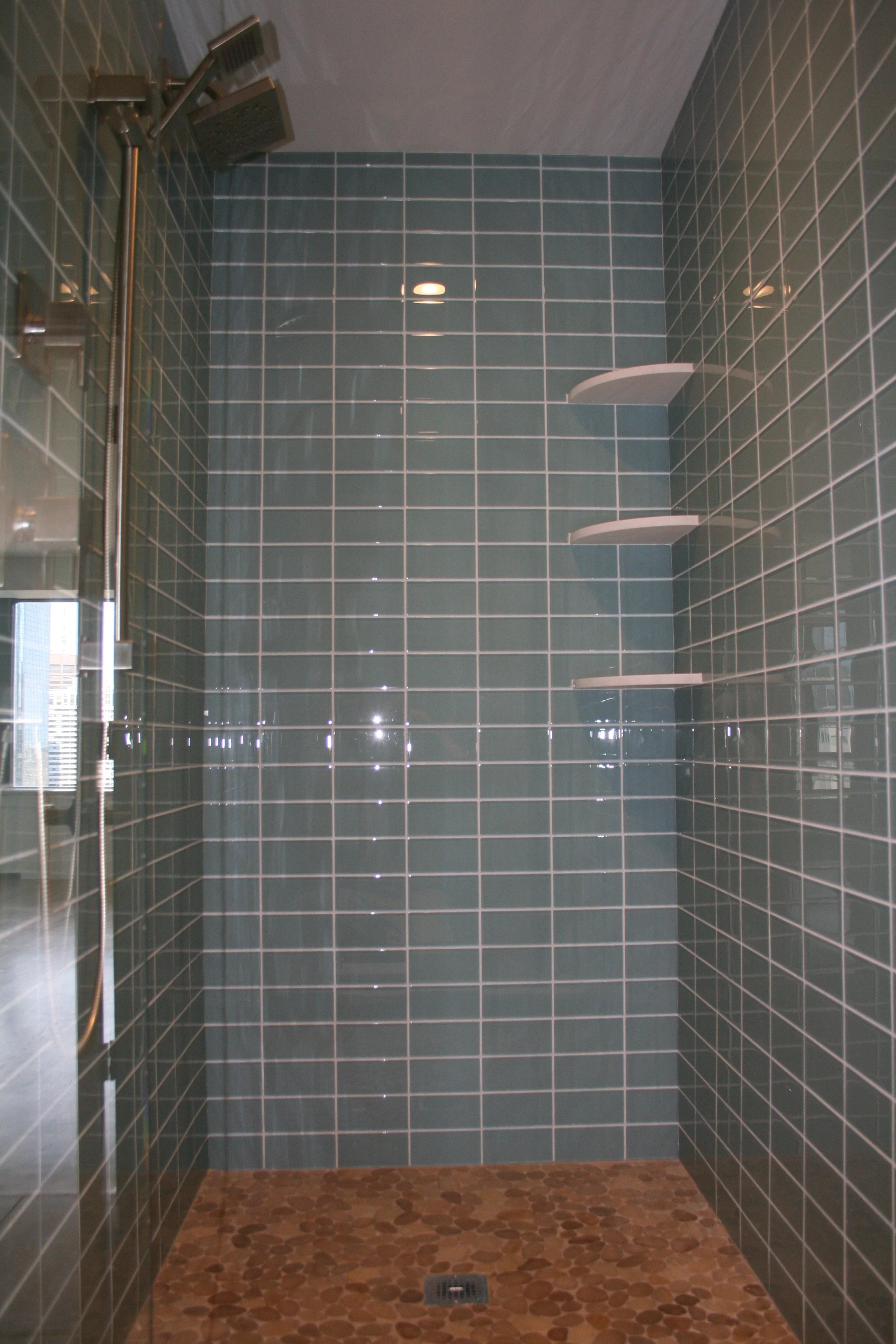 A modern shower with teal ceramic tiles on the walls, a rainfall showerhead, three white corner shelves, and a pebble stone floor with a drain.