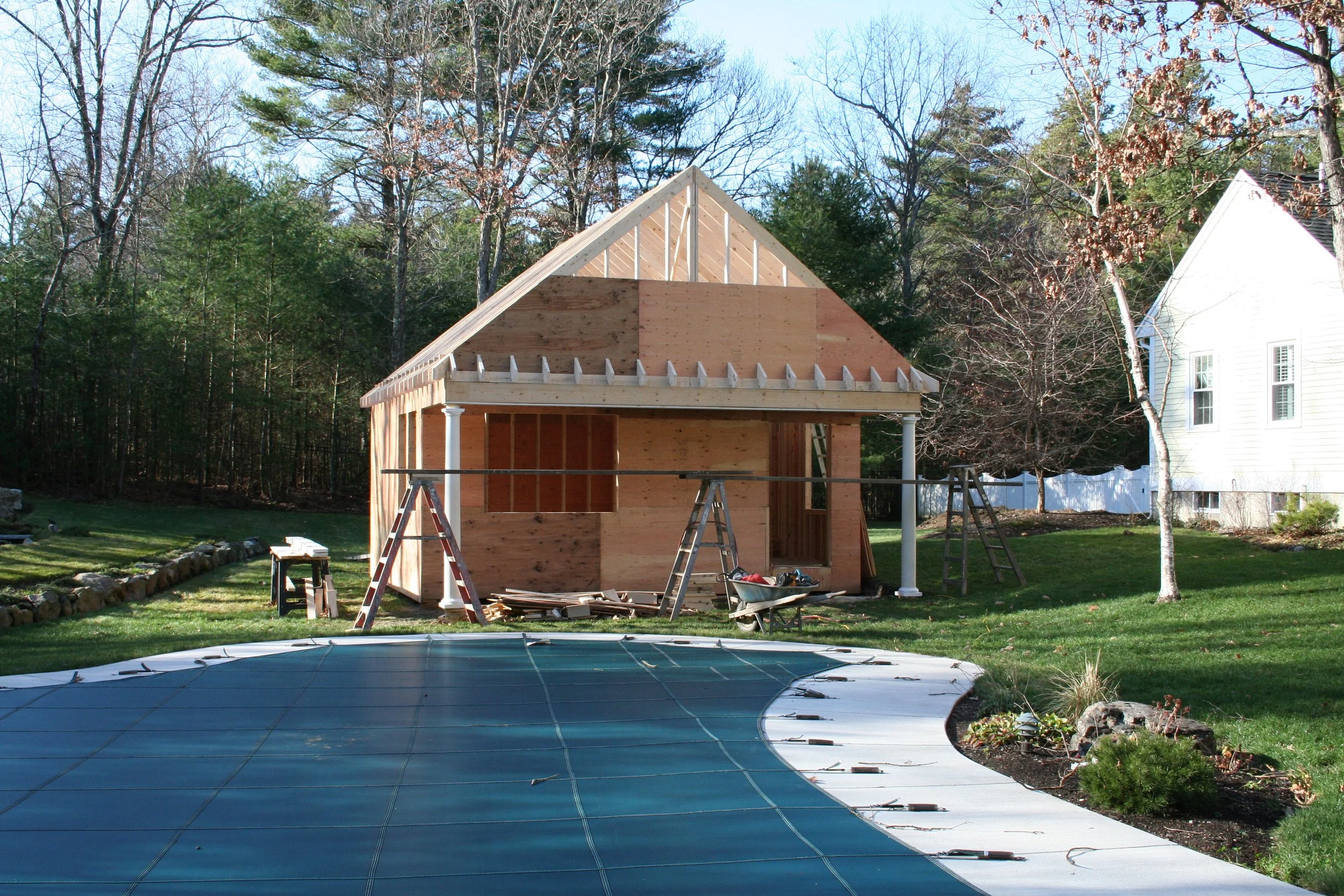 A house under construction with wooden framing and no roof. There is a ladder and construction tools in the yard. A swimming pool with a cover is in the foreground, surrounded by a lawn and trees.