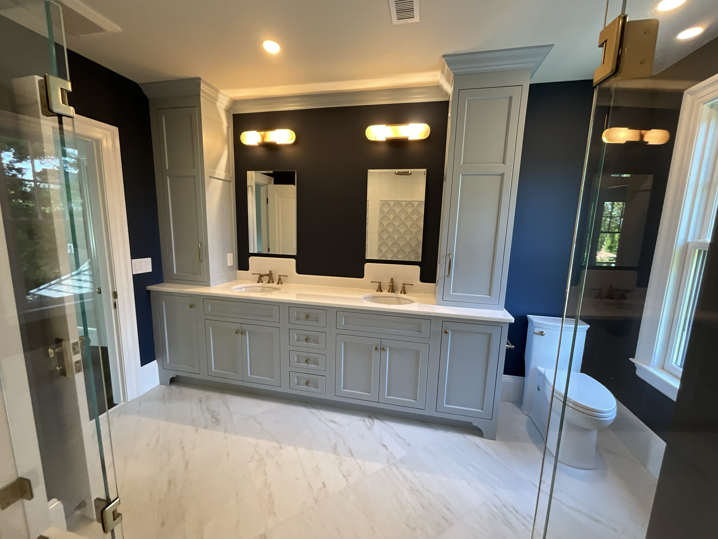 Modern bathroom with a double vanity, dark blue walls, white cabinetry, and marble flooring. There is a toilet in the corner and a glass shower enclosure on the left. The vanity has two mirrors and wall-mounted light fixtures above each mirror.
