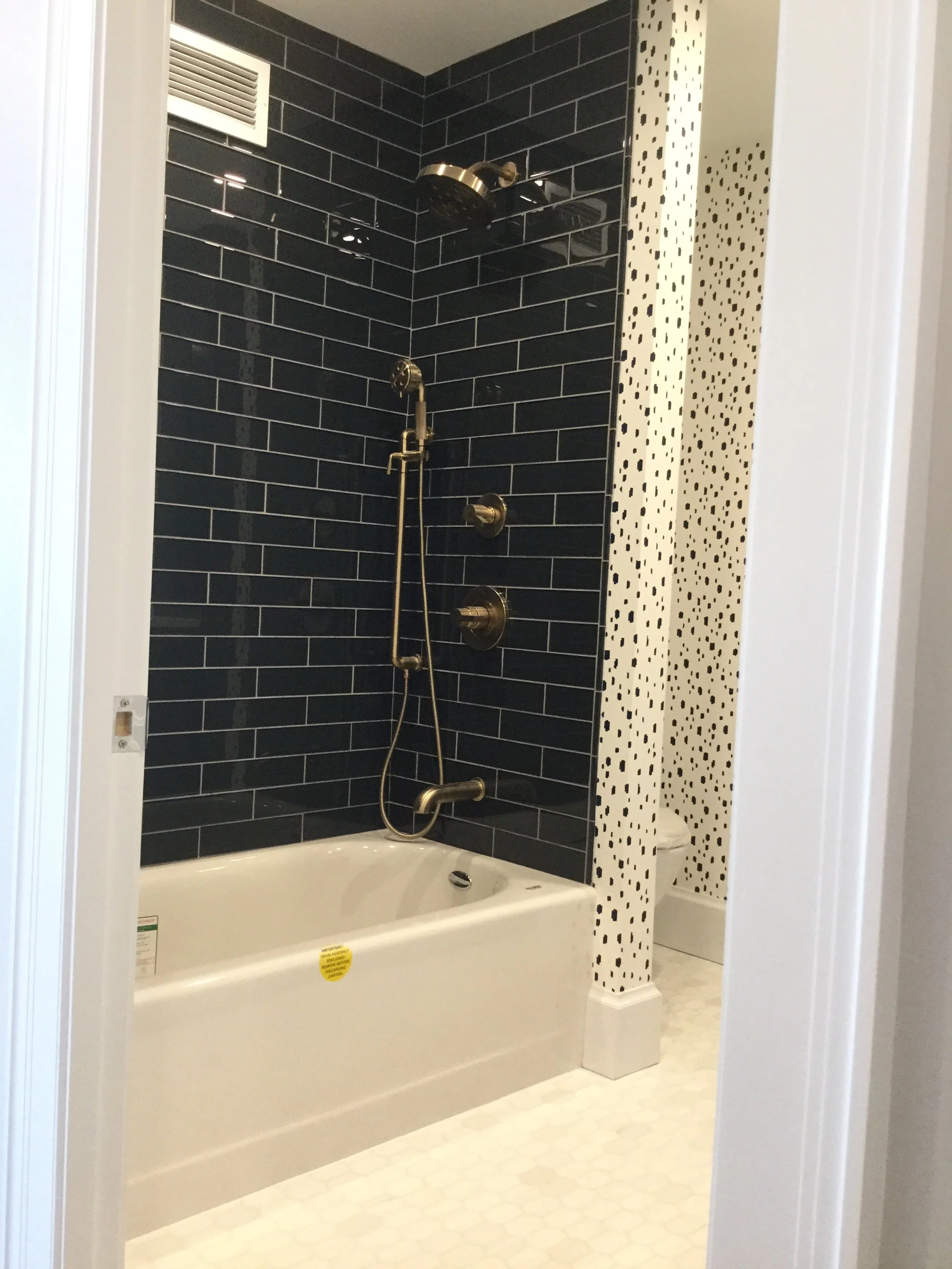 Bathroom shower with dark tile wall, vintage brass fixtures, and a white bathtub. Partial view of toilet behind a dotted accent wall.