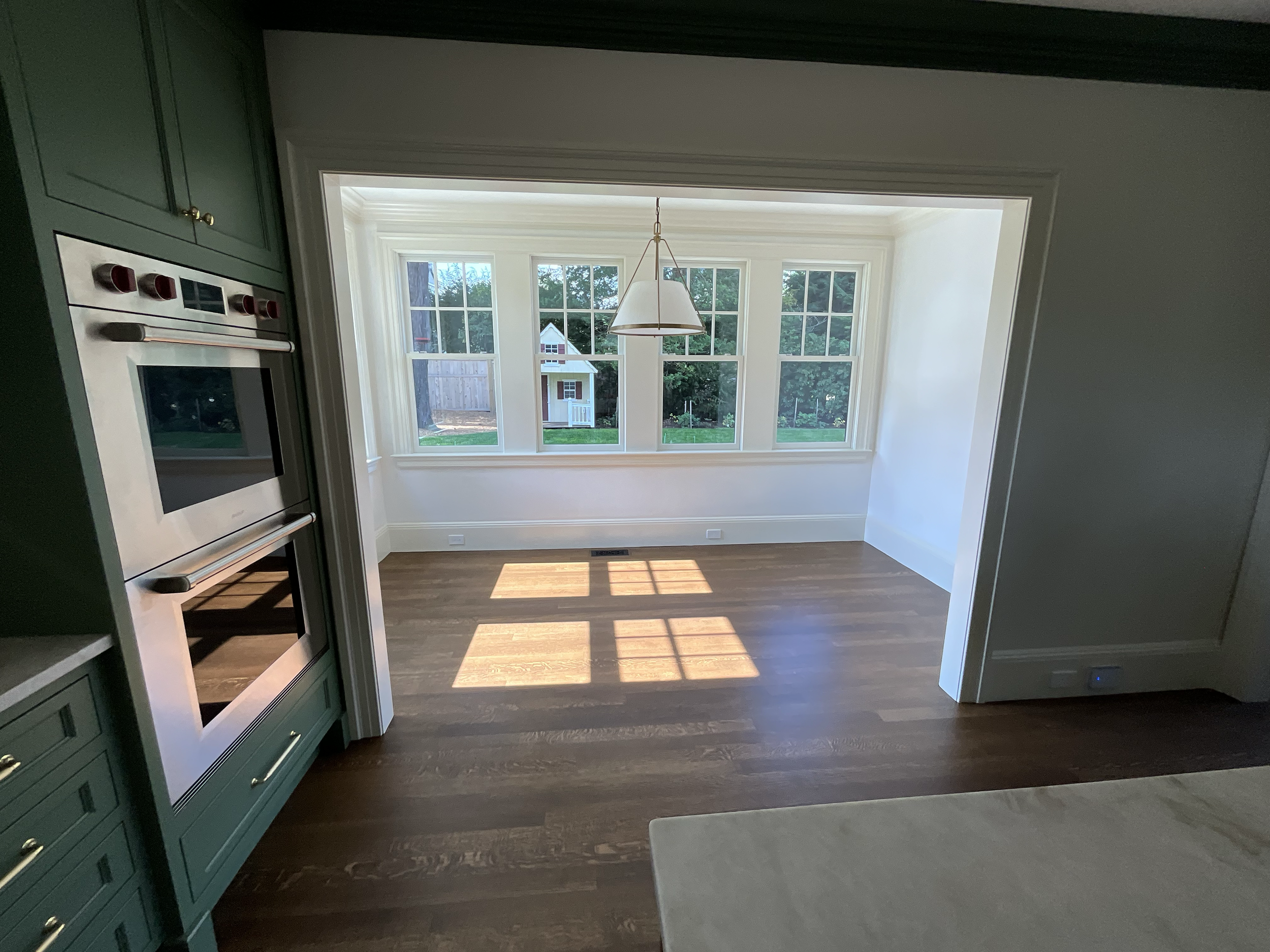 Bright room with large windows, hardwood floors, and a hanging white pendant light, viewed from the kitchen area.