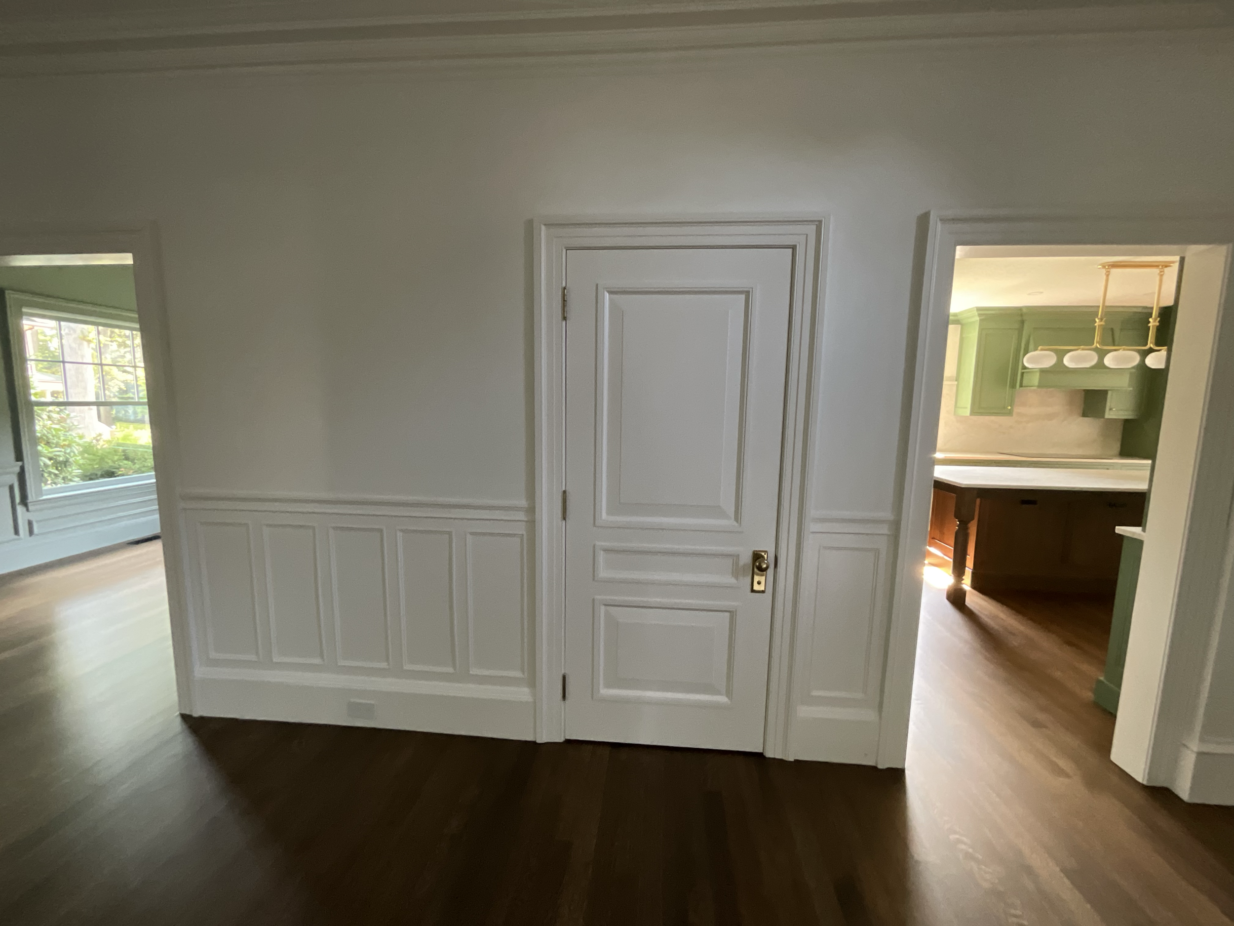 Interior view of a home with a closed white door, hardwood floors, and openings to a living room with large windows and a kitchen with green cabinets and a rustic wooden island.