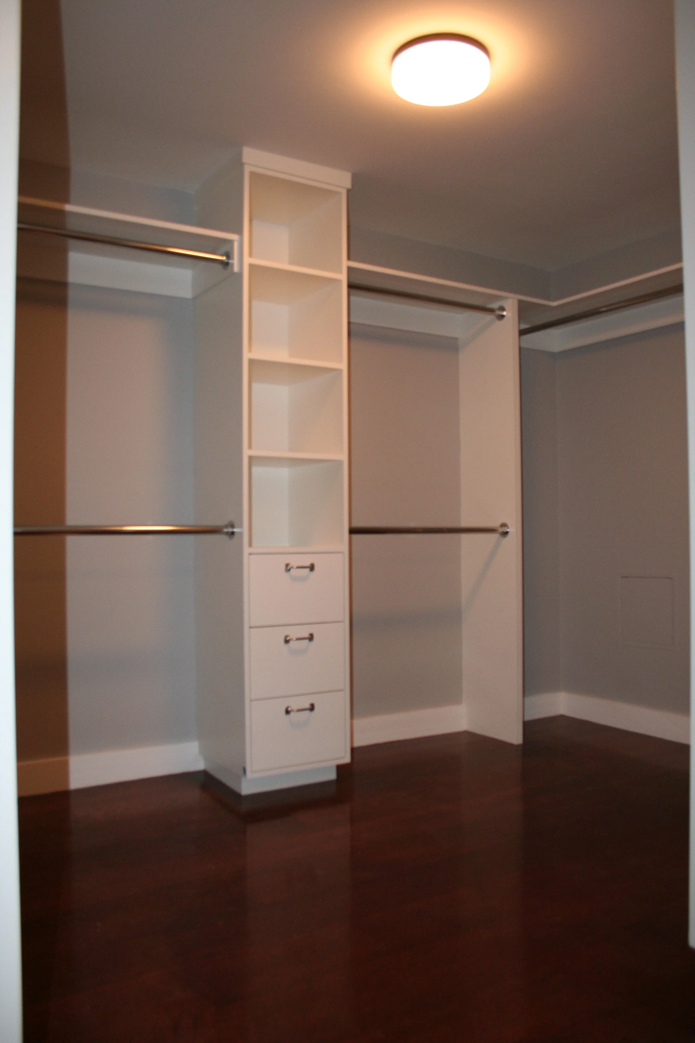Empty walk-in closet with white shelves, drawers, and hanging rods, illuminated by ceiling light, with dark hardwood floors.