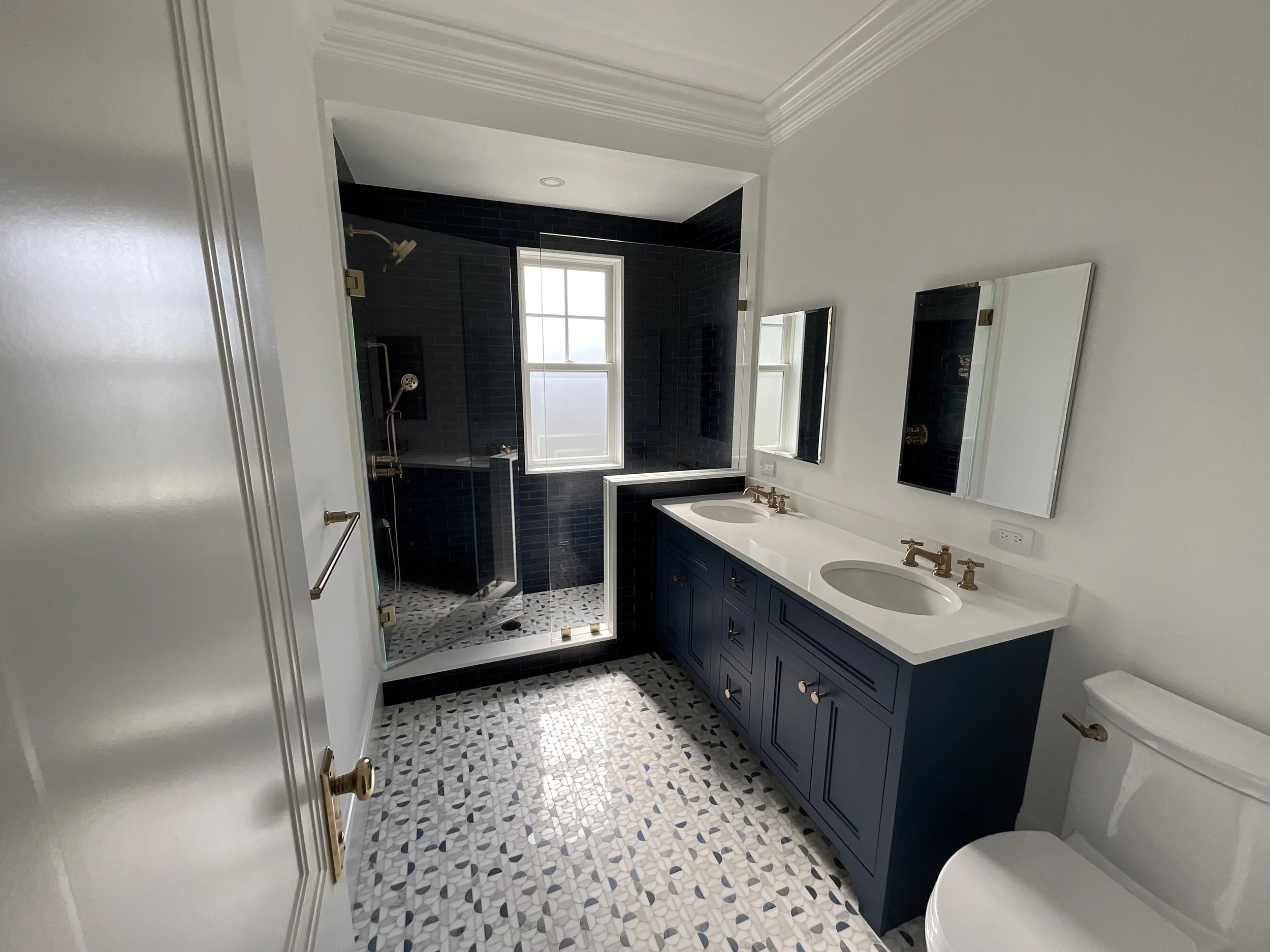 Modern bathroom with a double sink vanity, navy blue cabinets, white countertops, and two mirrors. There is a separate shower with dark tiles and a window, visible through an open doorway. The floor has a patterned tile design.
