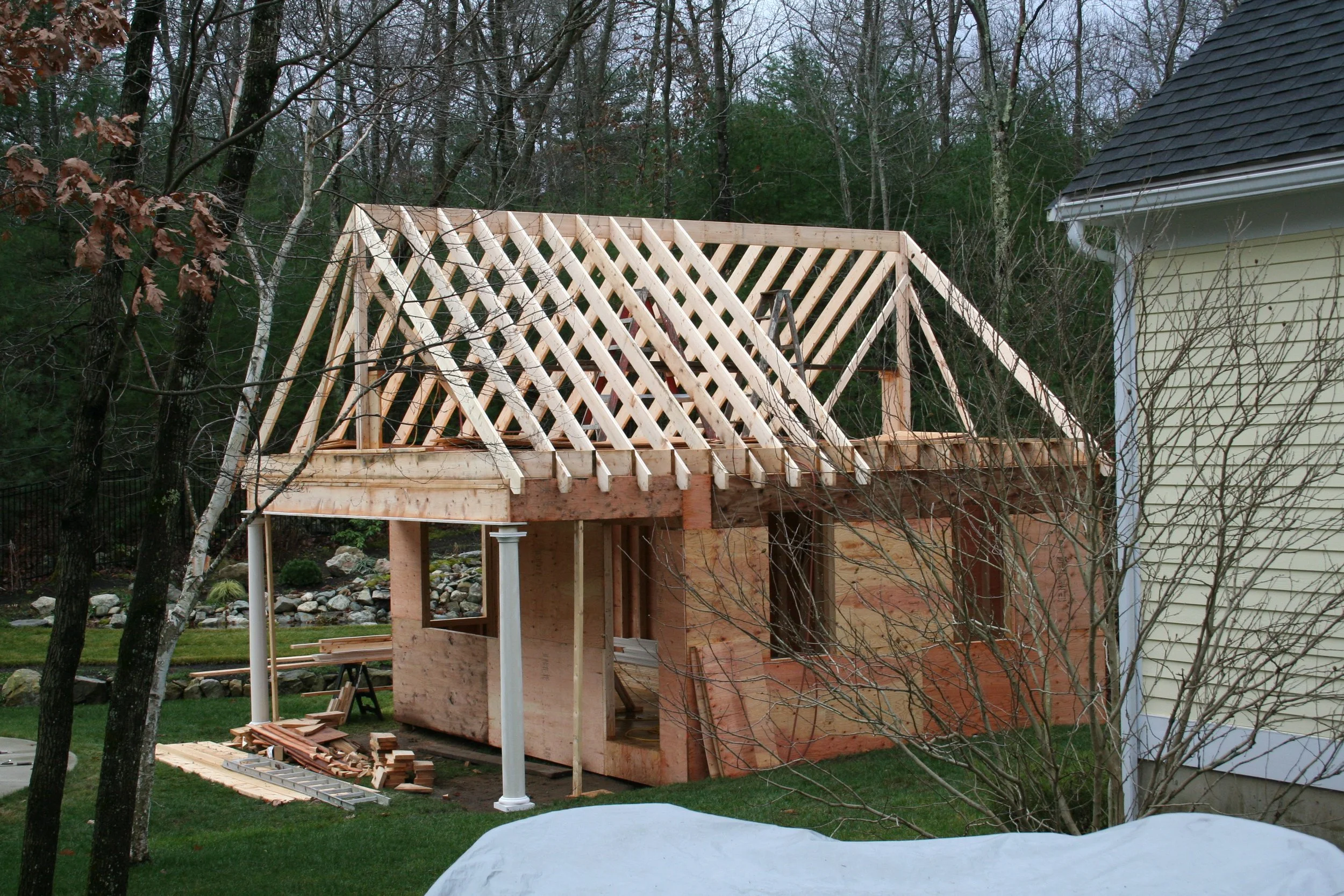 Construction of a wooden deck or porch extension on a residential house, showing framing and support columns, with building materials on the ground and trees in the background.