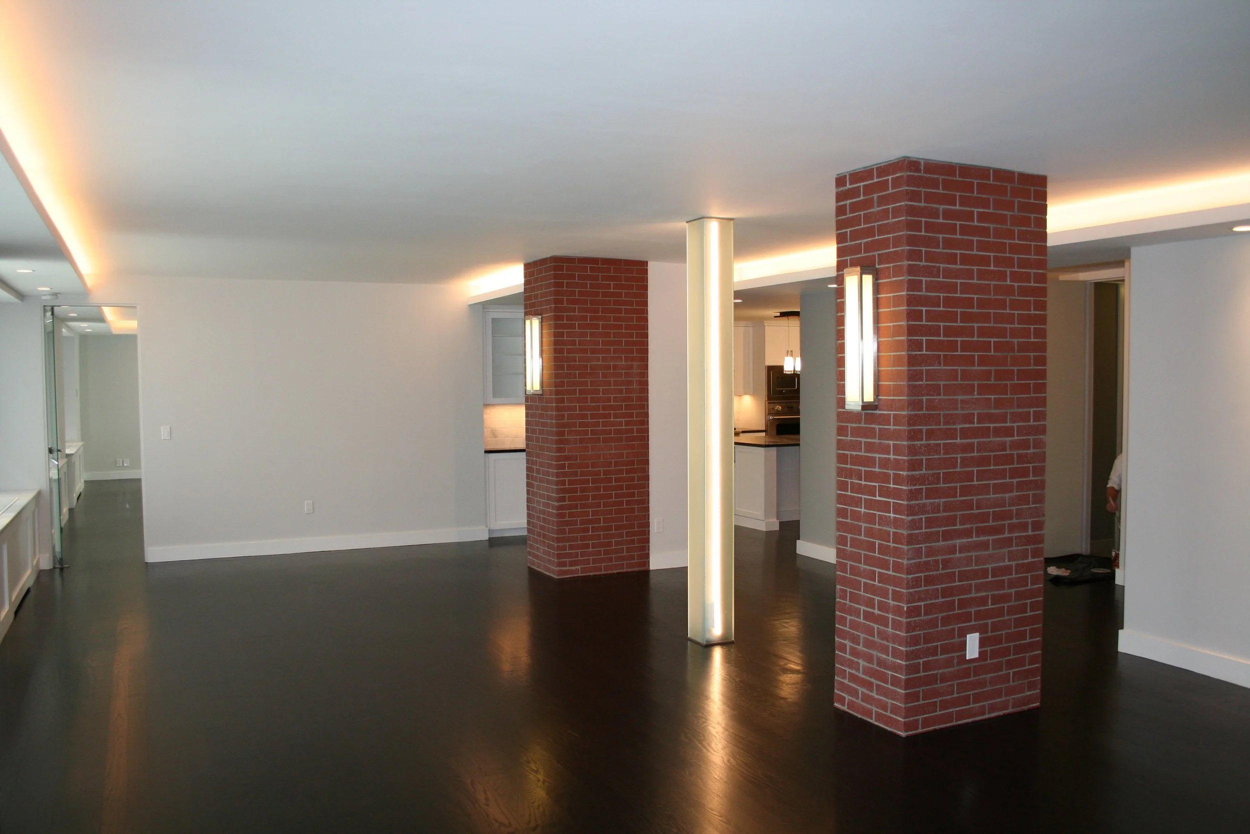 Empty modern apartment with dark wood floors, white walls, brick columns, and recessed lighting, with a view into the kitchen area.