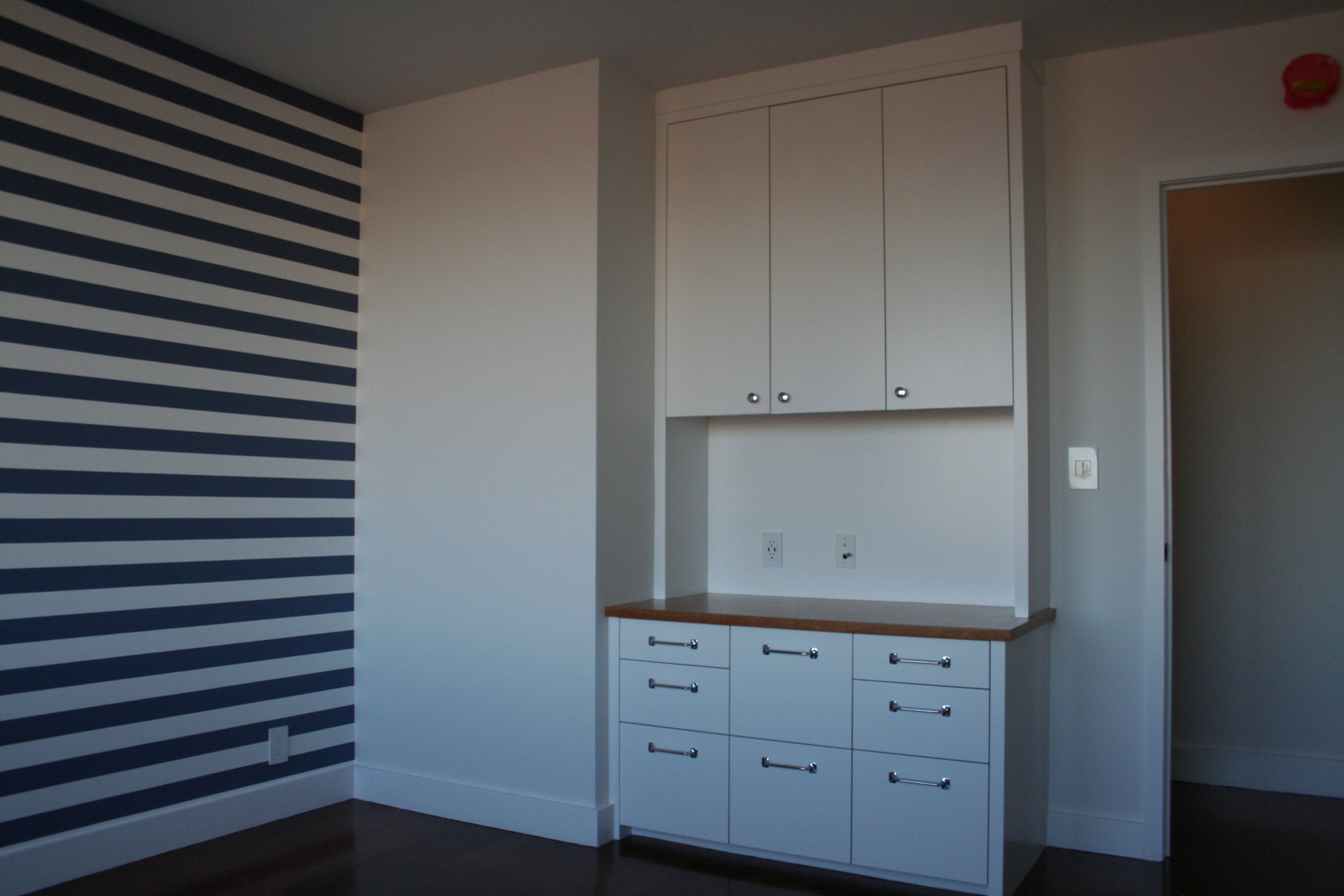 Interior of a room with a built-in white cabinet with multiple drawers and upper cabinets, a wooden countertop, a section of wall with navy blue and white horizontal stripes, and an electrical outlet and switches on the wall.