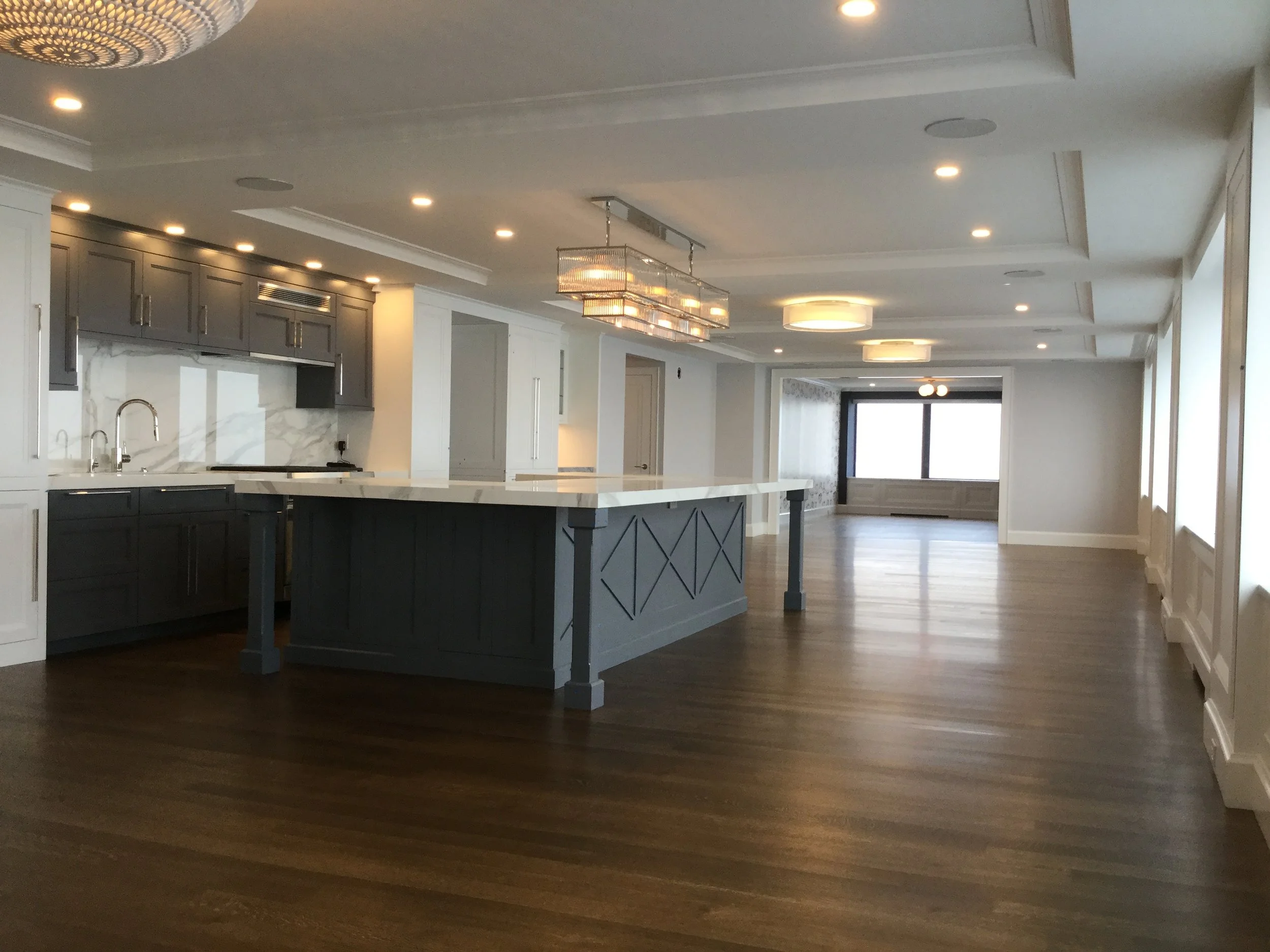 Empty, modern open-concept kitchen and living space with dark wood flooring, gray and white cabinetry, large island with white marble countertop, and ceiling lights.