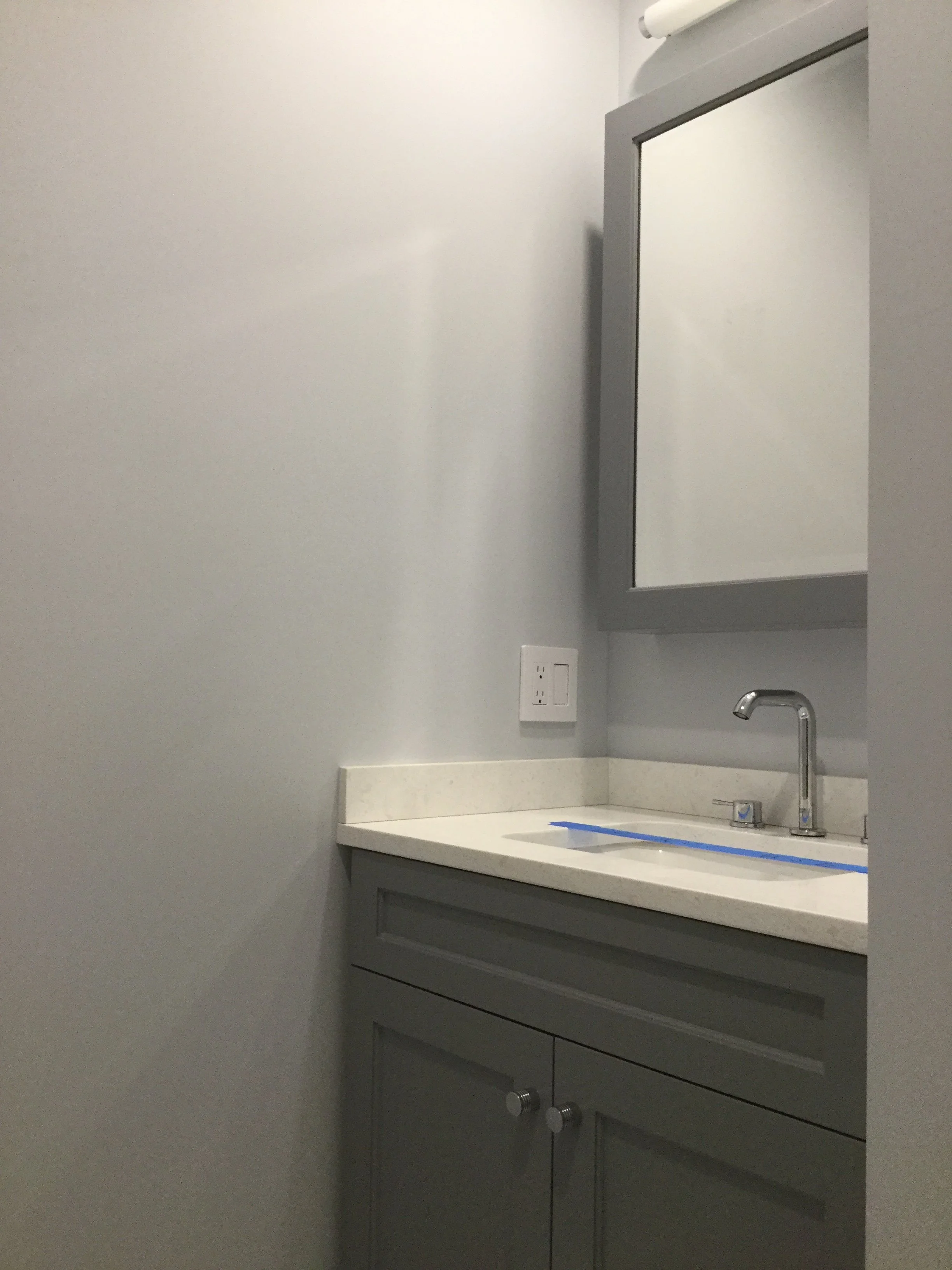 A bathroom vanity with a gray cabinet, a white countertop, a silver faucet, and a mirror above. An electrical outlet is on the wall next to the mirror.