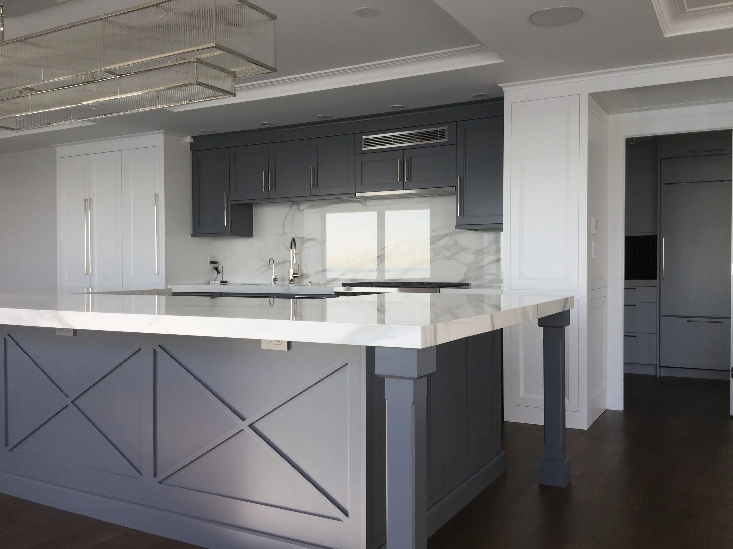 Modern kitchen with white marble countertops, dark gray lower and upper cabinets, a built-in oven, and a large island with a decorative front panel, set in a room with white walls and dark hardwood floors.
