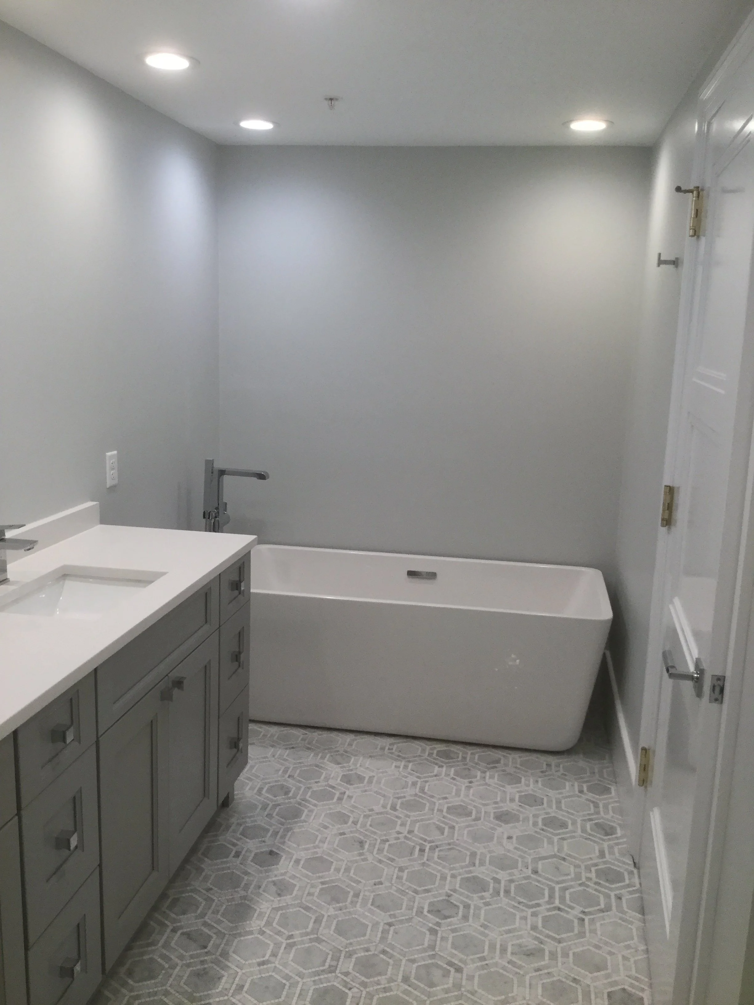 A modern bathroom with gray cabinetry, a white countertop with a sink, a bathtub, and gray patterned floor tiles, illuminated by recessed ceiling lights.