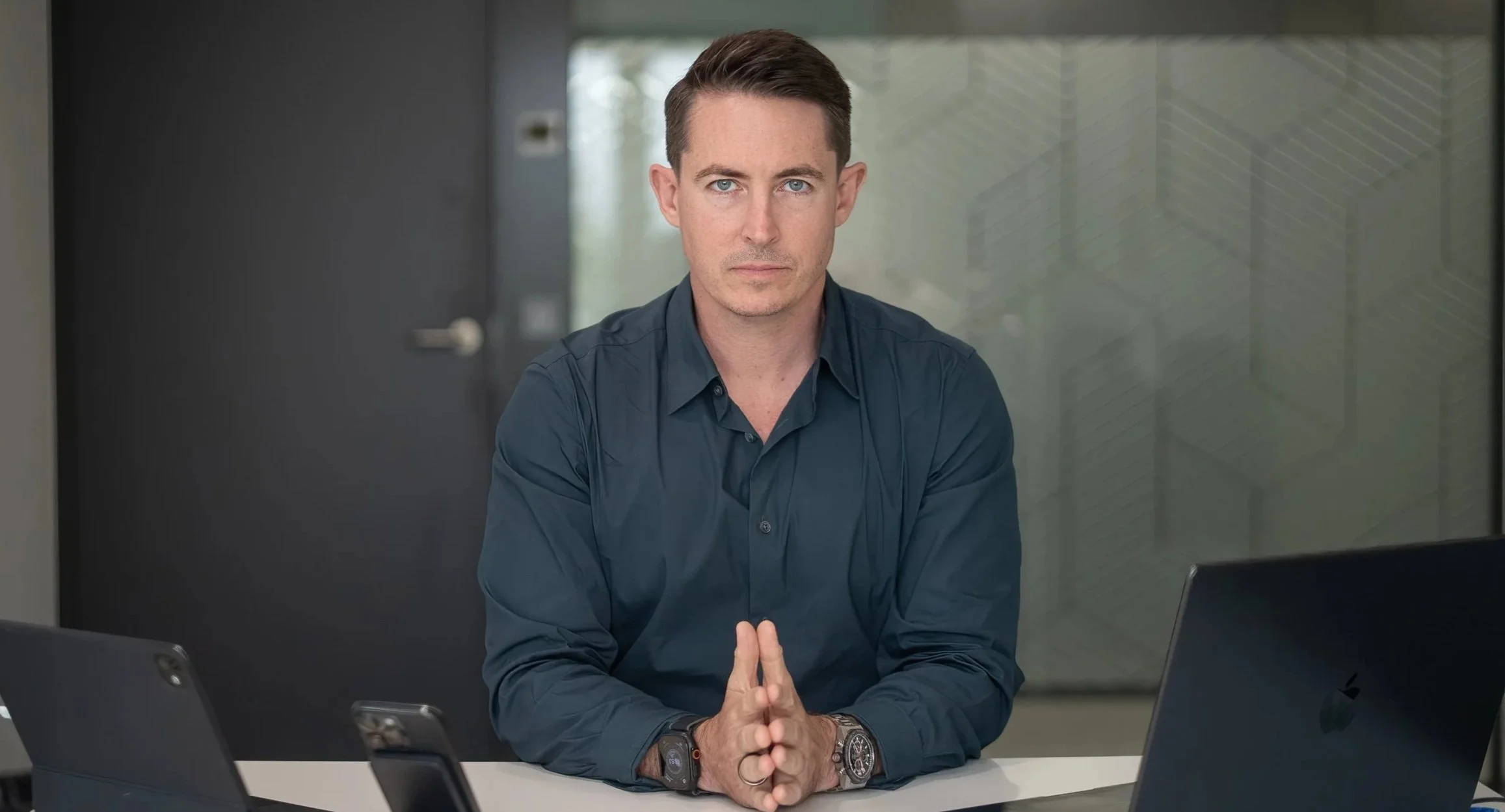 A man with short dark hair and blue eyes sitting at a desk with his hands clasped together, wearing a dark blue button-up shirt and a wristwatch, looking directly at the camera in a modern office.