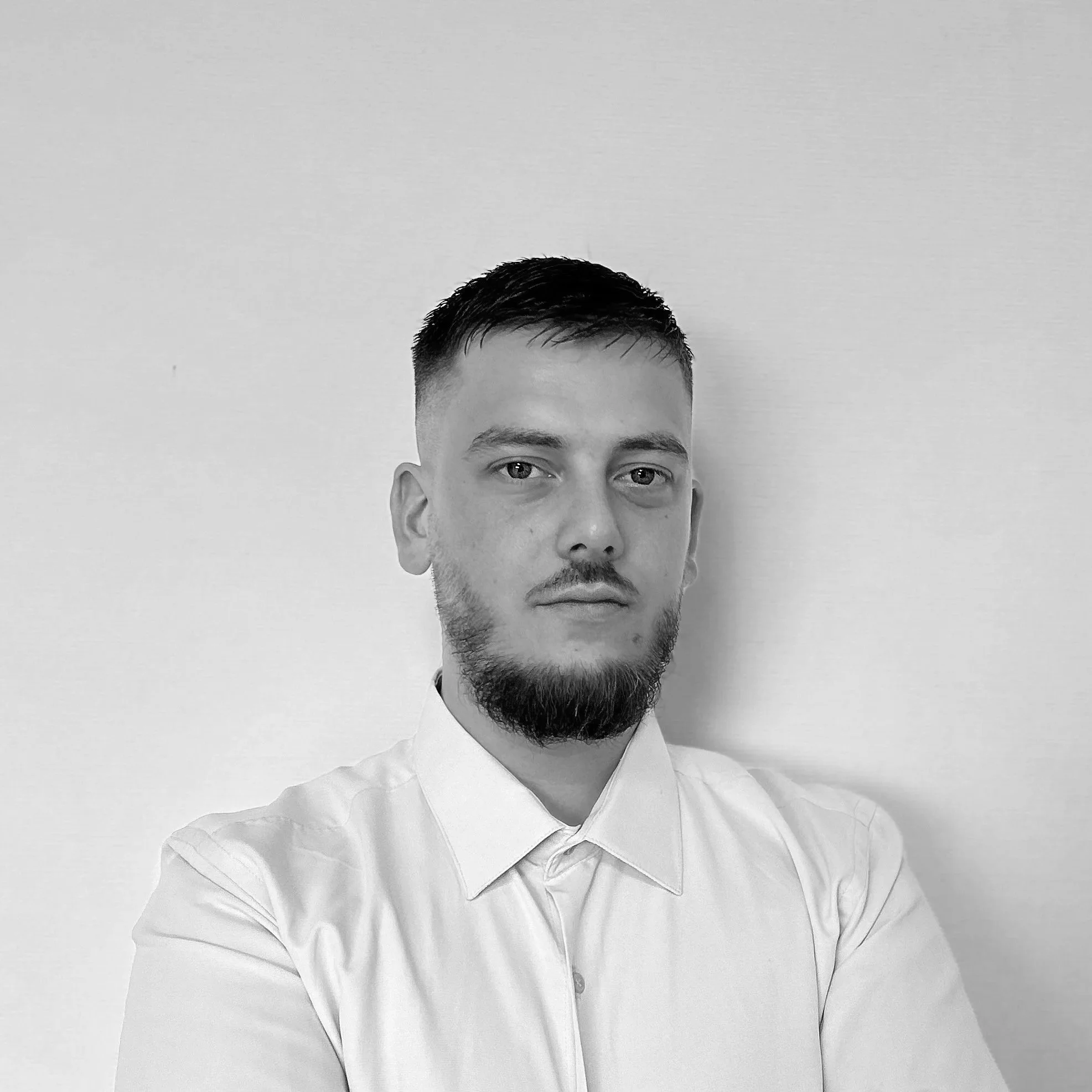 Portrait en noir et blanc d'un homme avec une barbe, portant une chemise blanche, regardant vers l'avant.