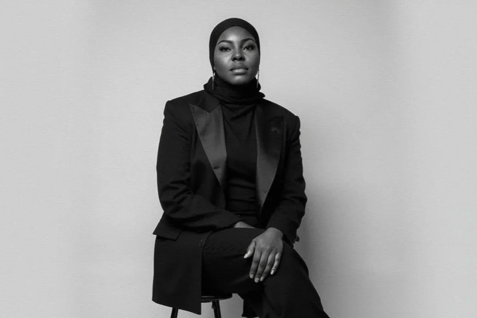 Femme noire habillée en noir, portant un hijab, assise sur une chaise contre un mur blanc, en mode noir et blanc.