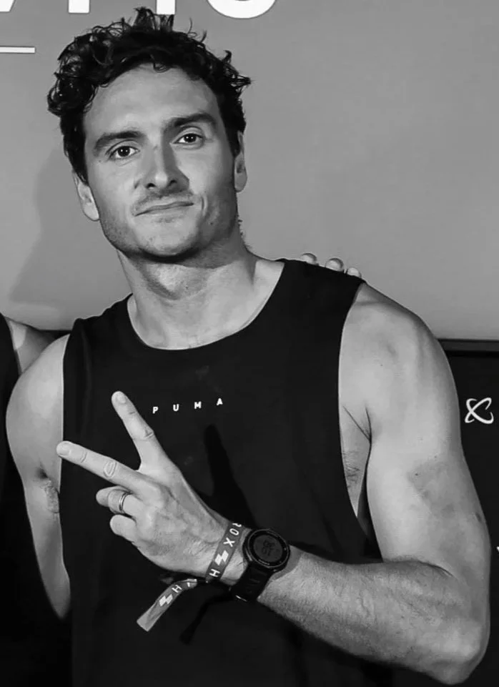 Homme avec cheveux bouclés, portant un débardeur noir, faisant un signe de paix avec la main droite, porte une montre et un bracelet