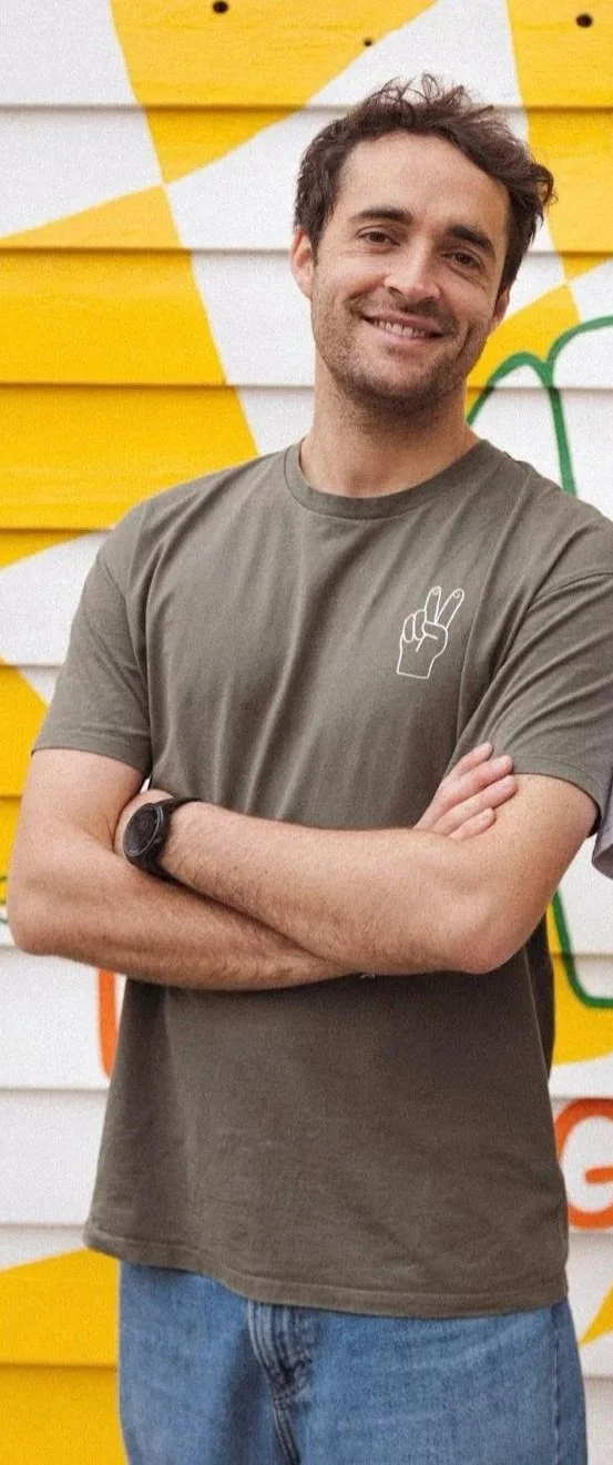 Un homme souriant avec les bras croisés, portant un t-shirt gris à une petite illustration de main faisant le signe de la paix, devant un mur peint jaune et blanc.