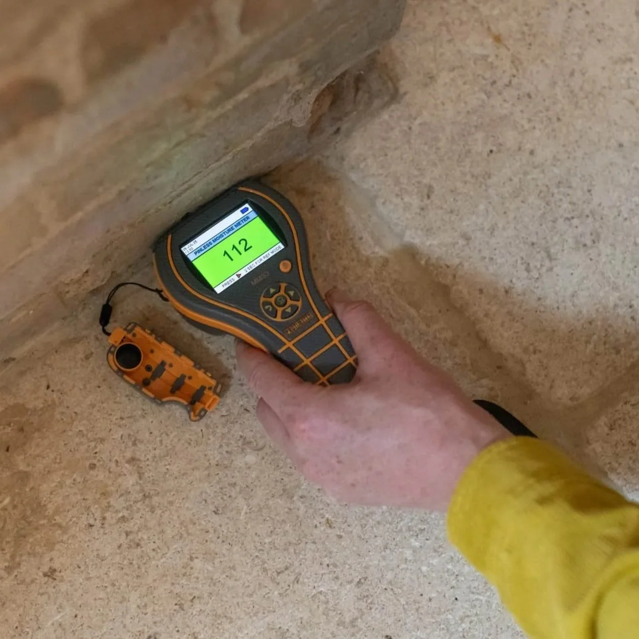 A person measuring moisture level in a rock wall using a moisture meter with a reading of 112.