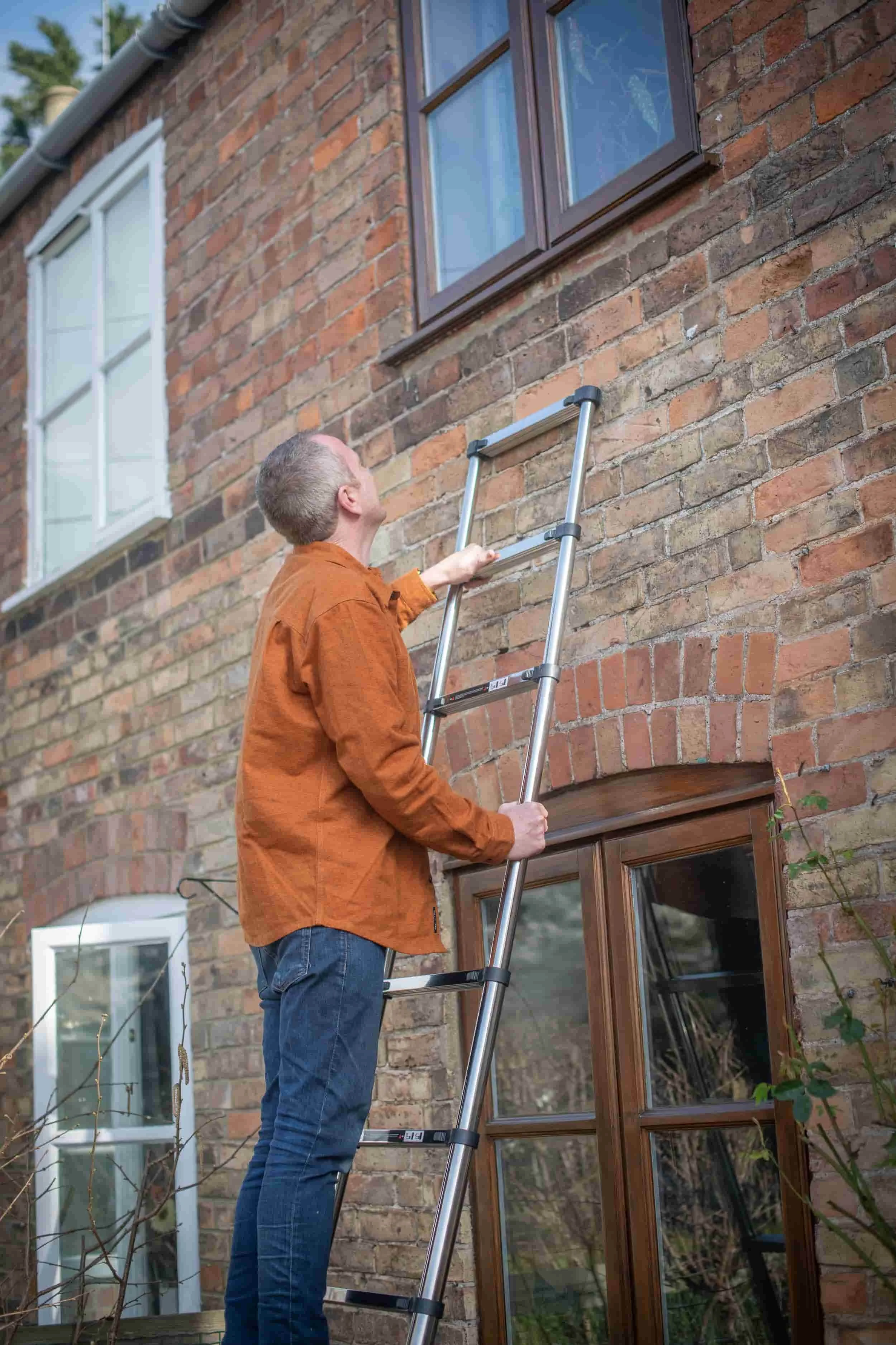 A man standing on an extension ladder outside a brick house, reaching up to clean or inspect a window.