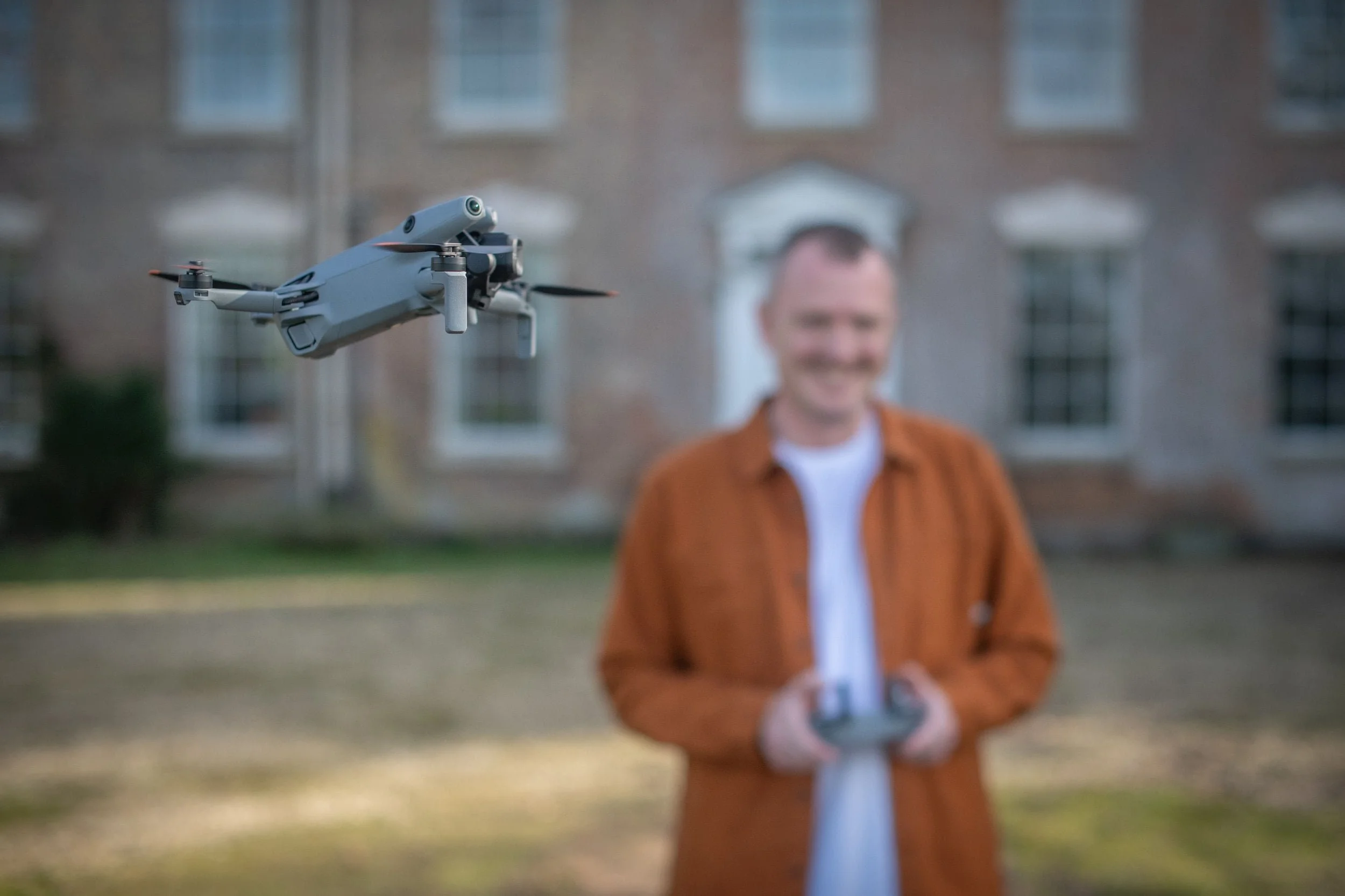 A drone flying in front of a smiling man holding a remote control in an outdoor setting with a blurred building in the background.
