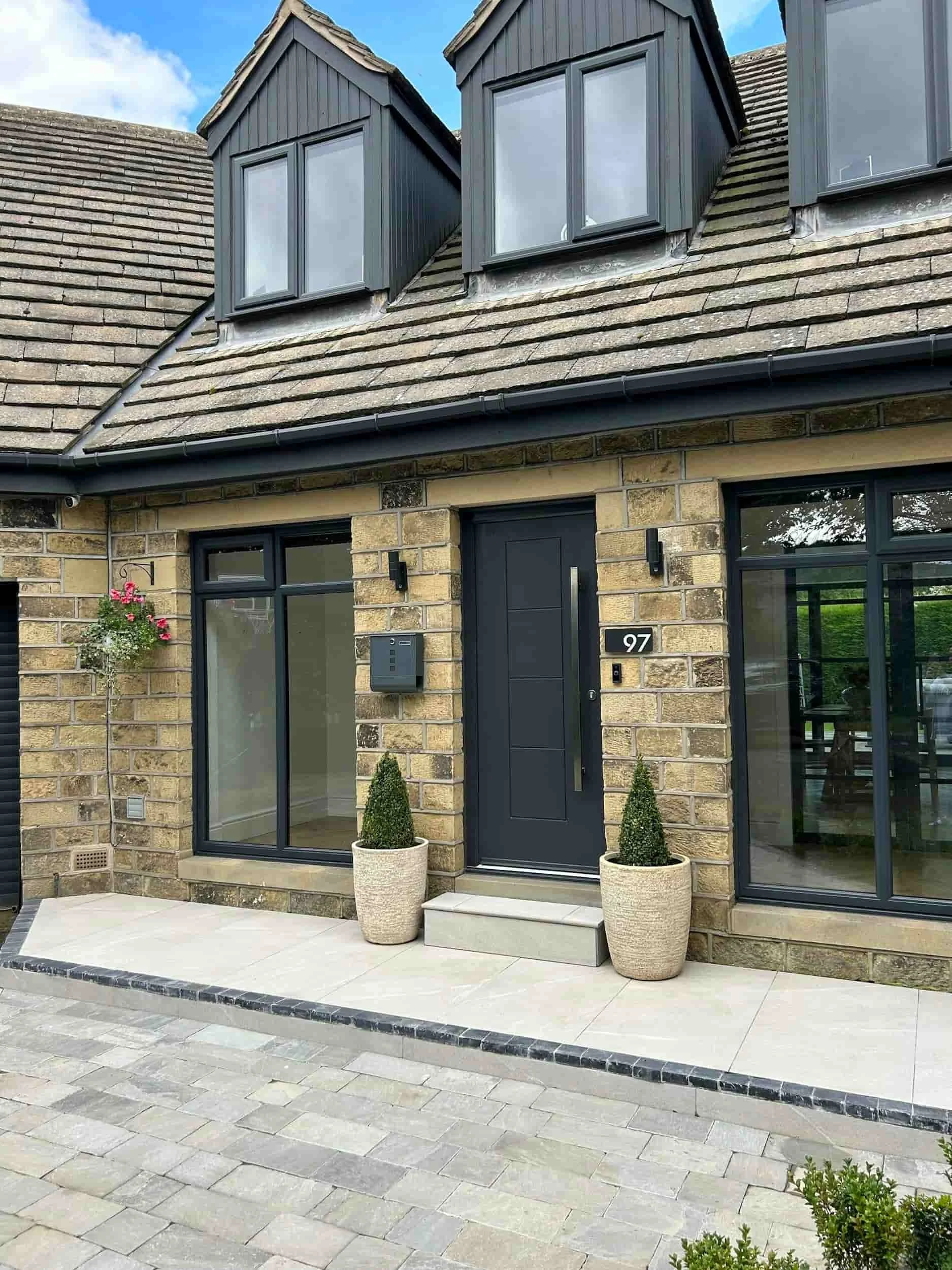 Front entrance of a modern house with a black door, large window, potted plants, and brick exterior.