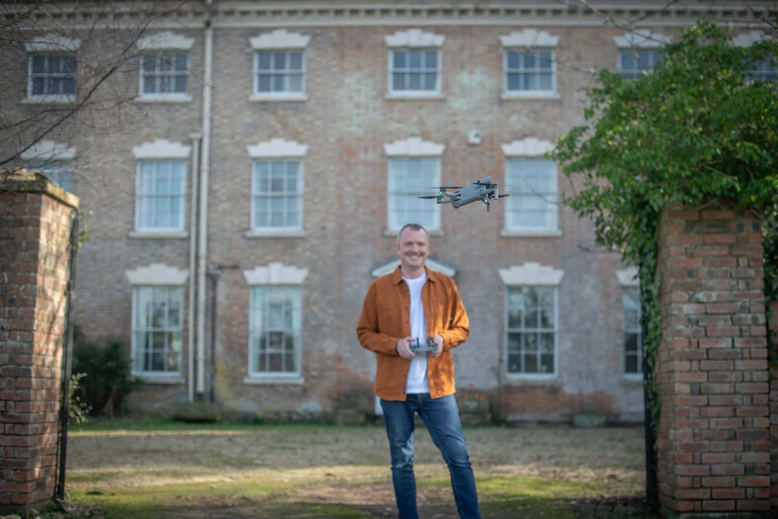 A man in an orange jacket flying a drone outdoors in front of a brick building.