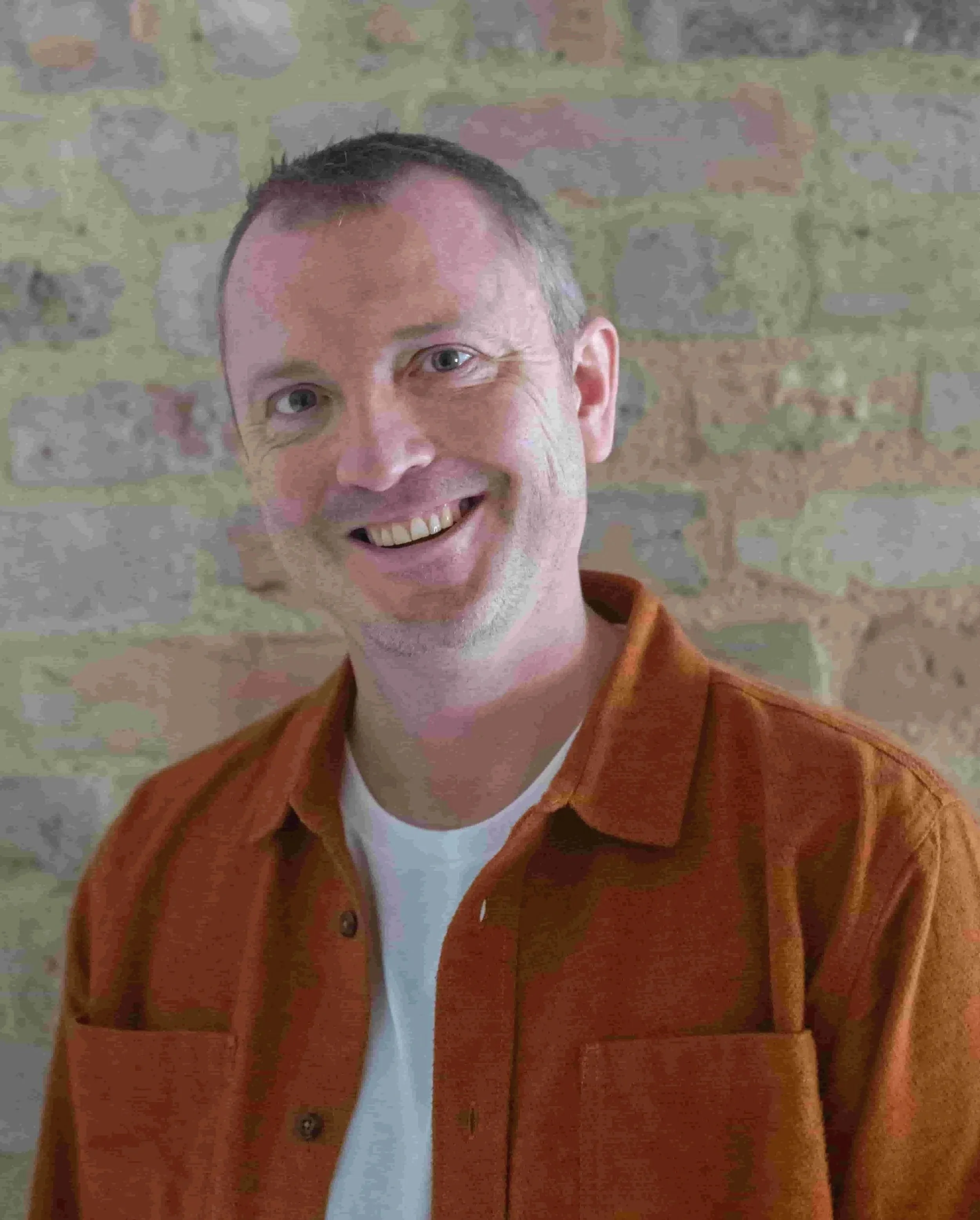 A smiling man with short brown hair and blue eyes, wearing an orange jacket over a white shirt, standing against a light brick wall.