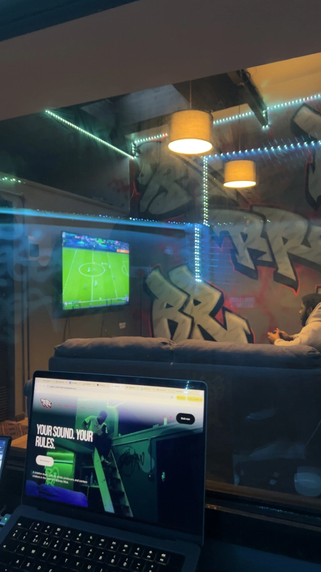 A person playing video games on a TV in a room with graffiti art on the wall, while another person is working on a laptop with the text "Your Sound. Your Rules." on the screen, reflecting an indoor gaming lounge environment.