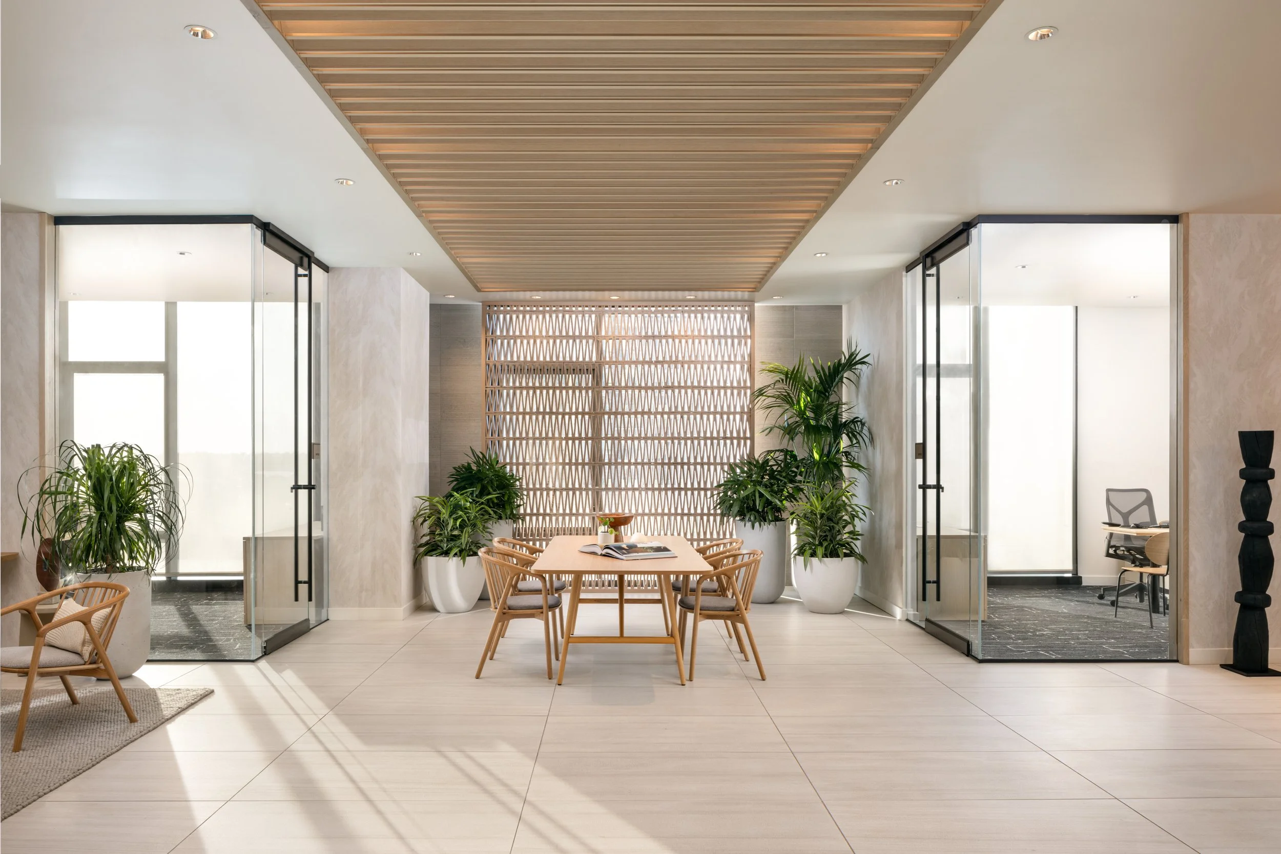 Modern conference room or office space with a central wooden table, six chairs, potted plants, glass doors, and a decorative wooden wall panel.